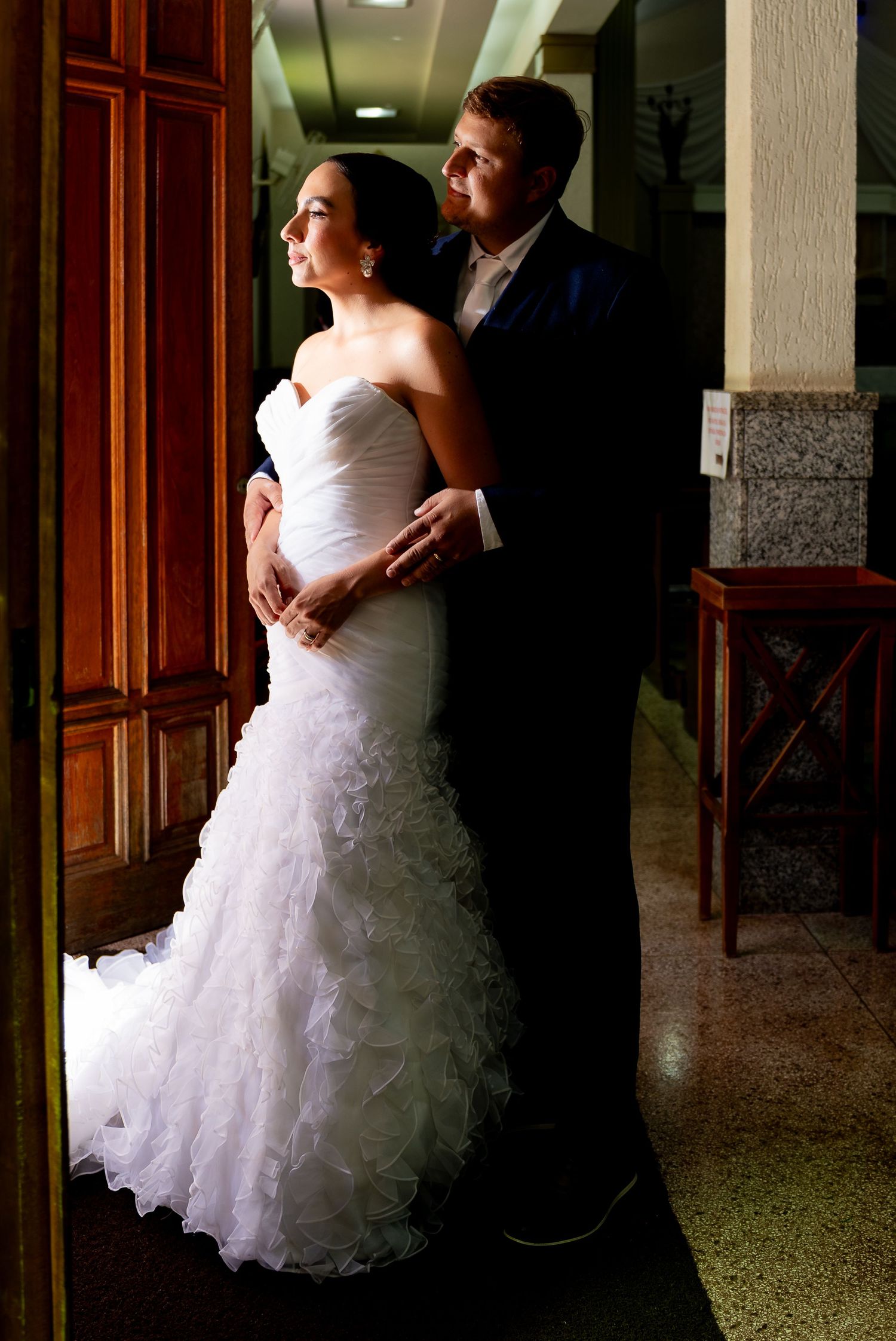 Bride in white ruffled gown and groom in navy suit share a tender embrace near a wooden door.