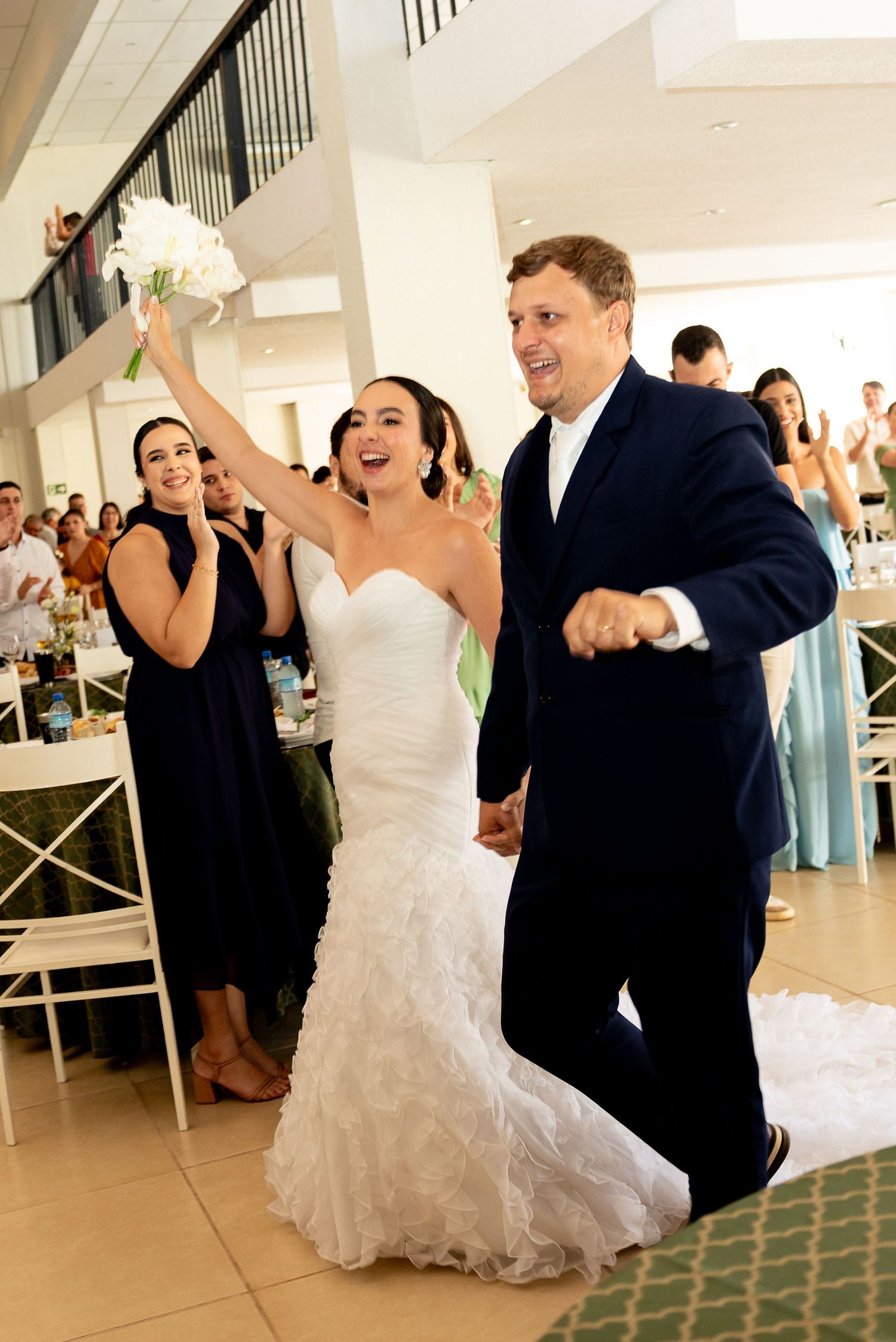 Joyful bride in white mermaid gown raises bouquet while groom in navy suit enters wedding reception.