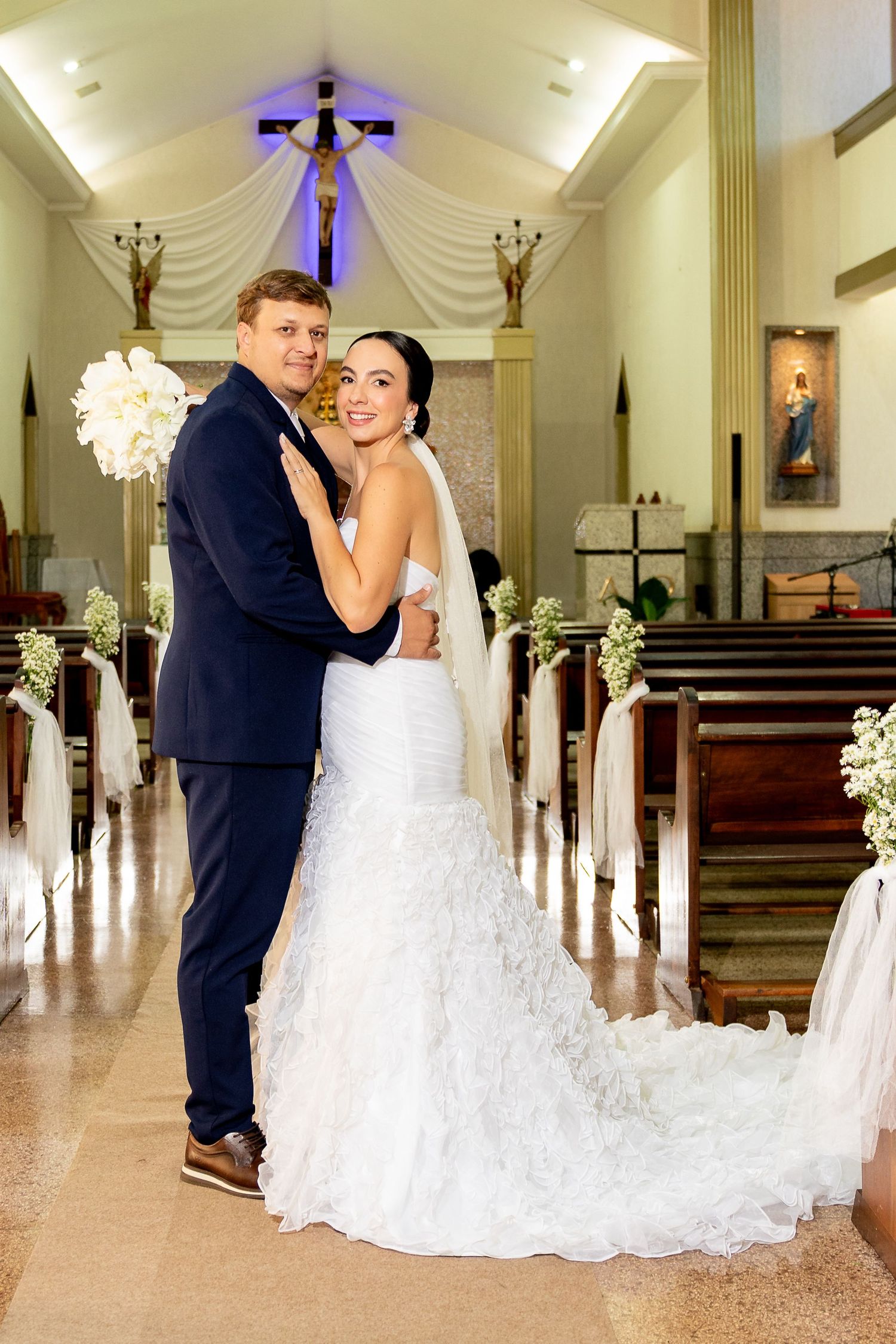 A bride and groom embrace at the altar of a decorated church, with the bride in a white mermaid gown holding a bouquet.