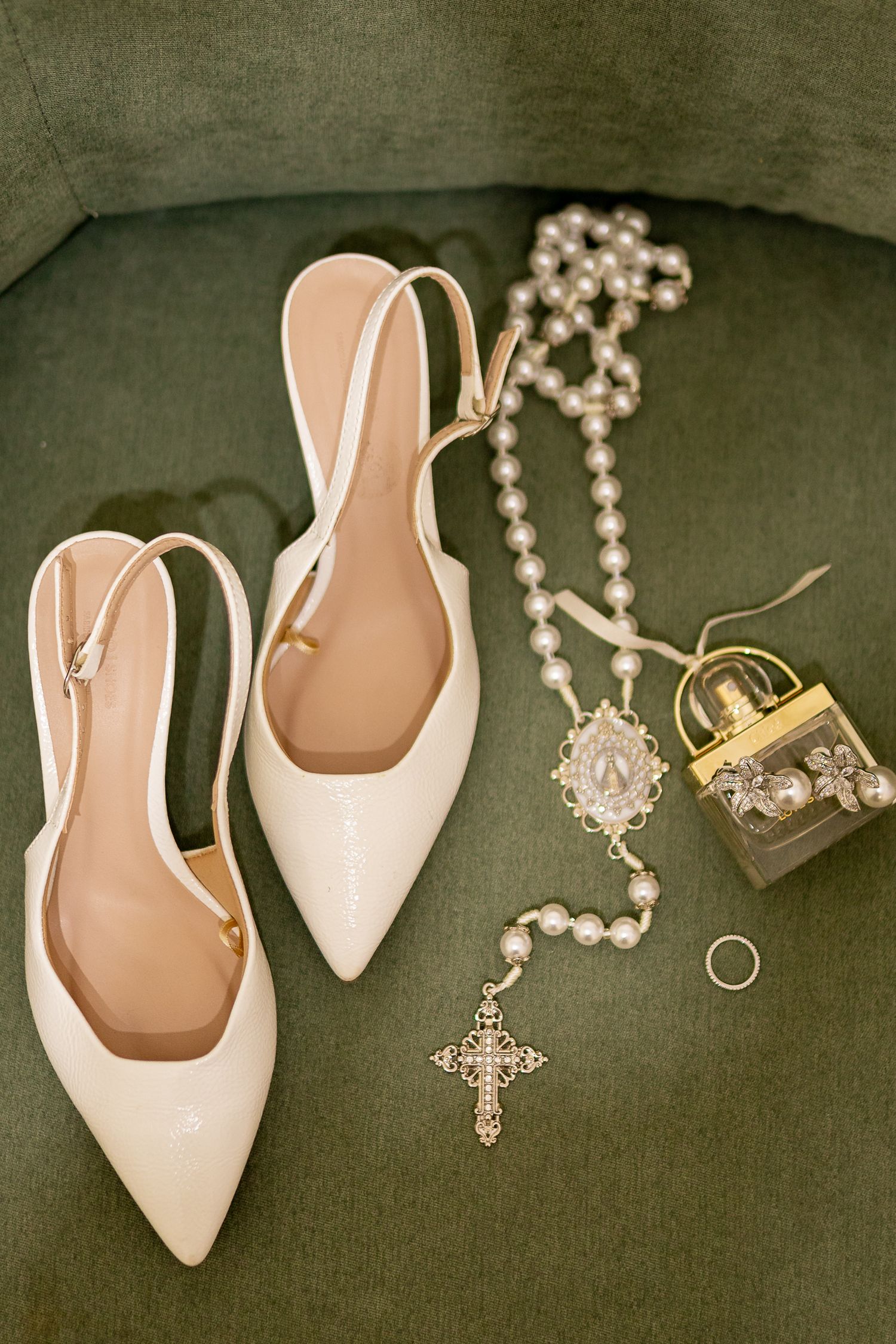 White slingback bridal heels displayed with pearl rosary, gold cross pendant, wedding ring, and ornate gold lock on green velvet.