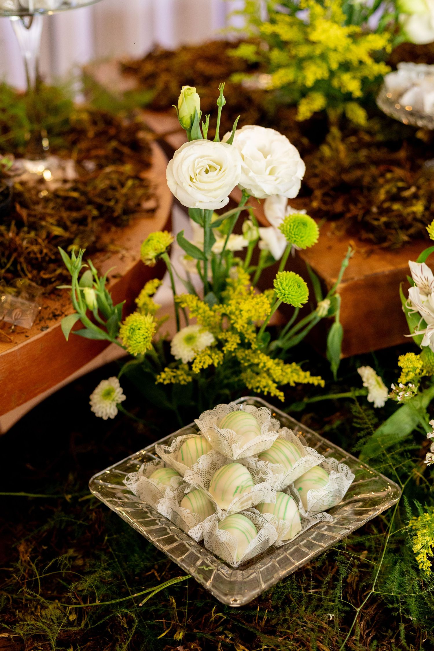 Elegant wedding dessert table with white ranunculus flowers and green-wrapped cake pops on a crystal plate.