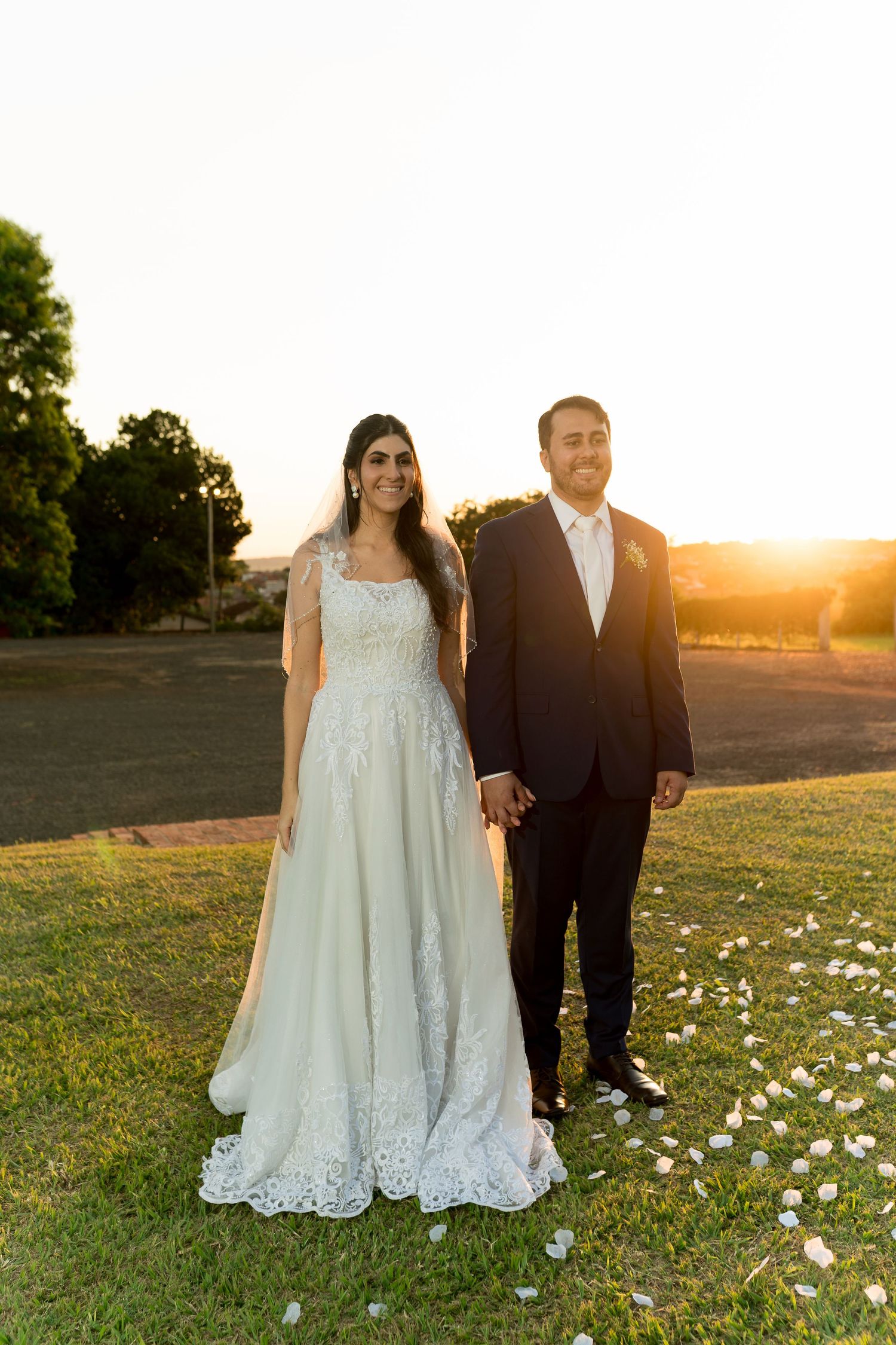 Bride and groom holding hands at sunset on green grass, bride in lace wedding gown, groom in black suit.