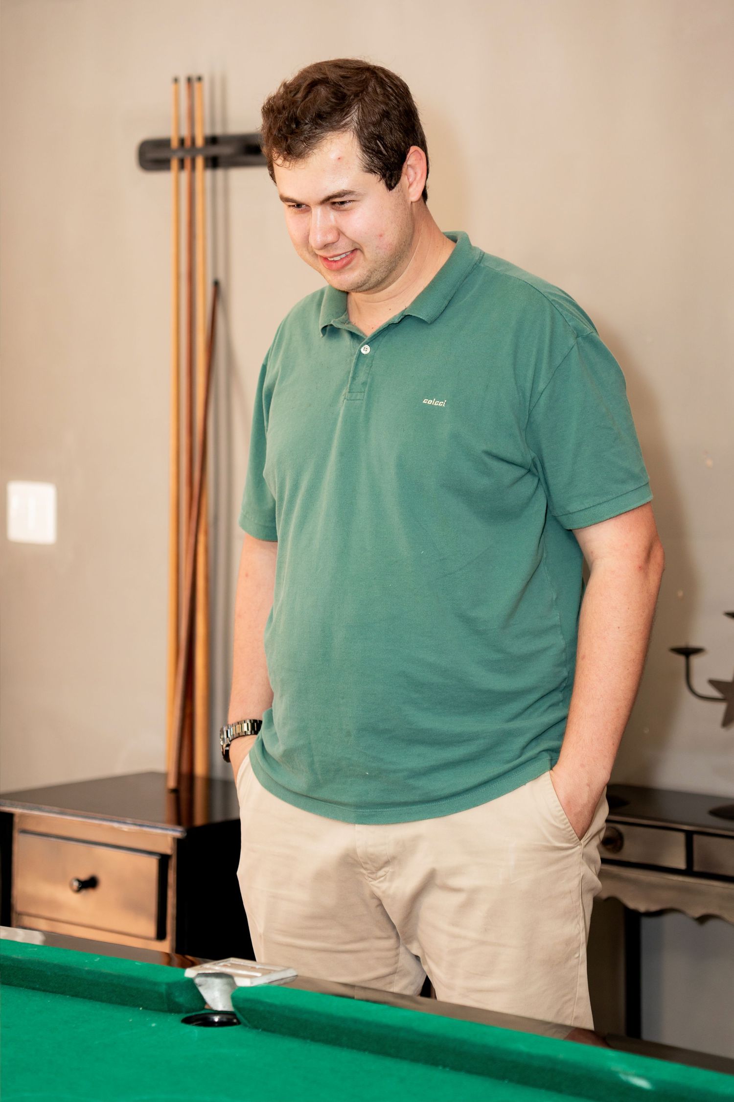 Man in green polo shirt standing by a pool table in a game room with billiard cues on the wall.
