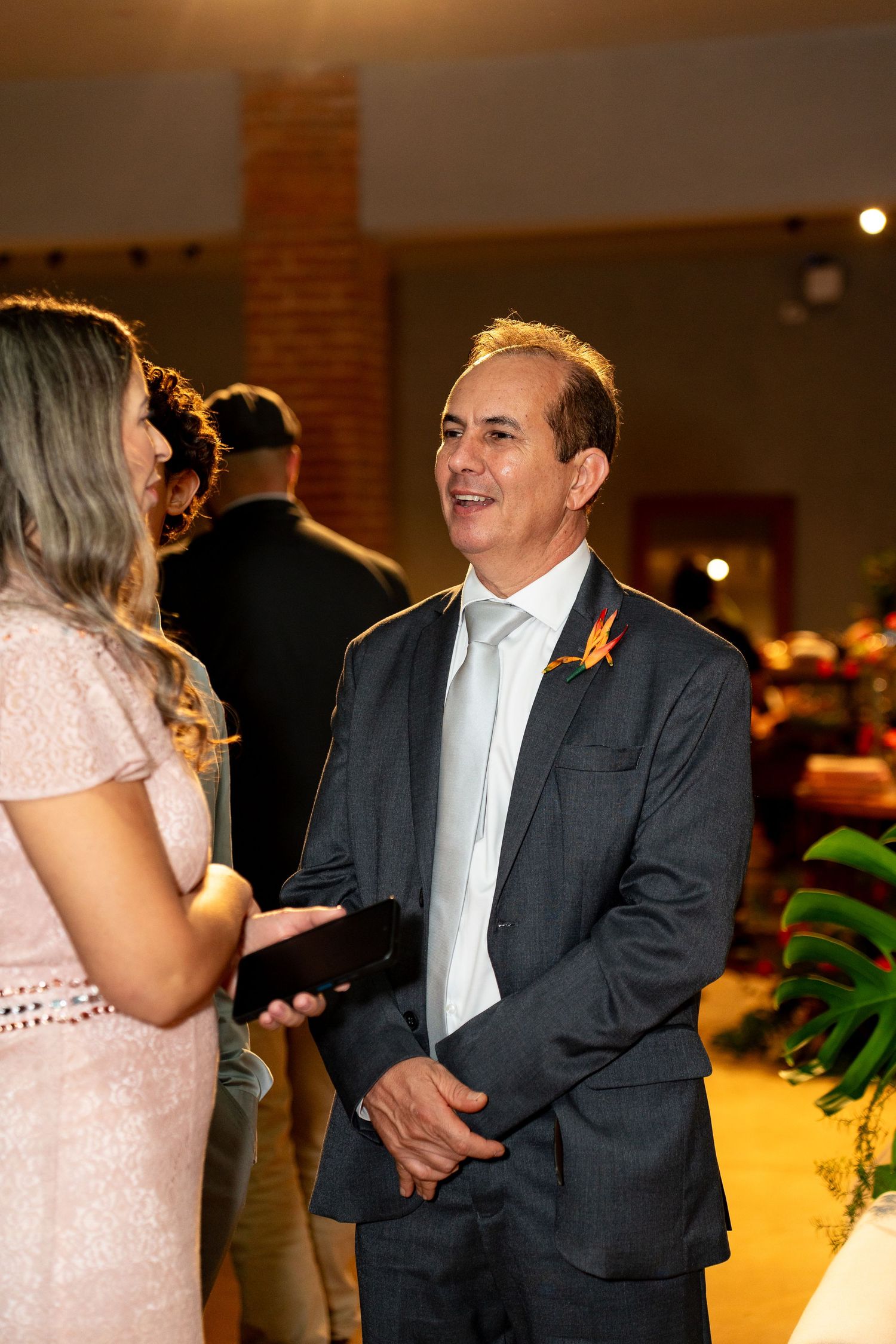 Man in dark suit with orange boutonniere smiling at woman in pink dress at formal indoor event.