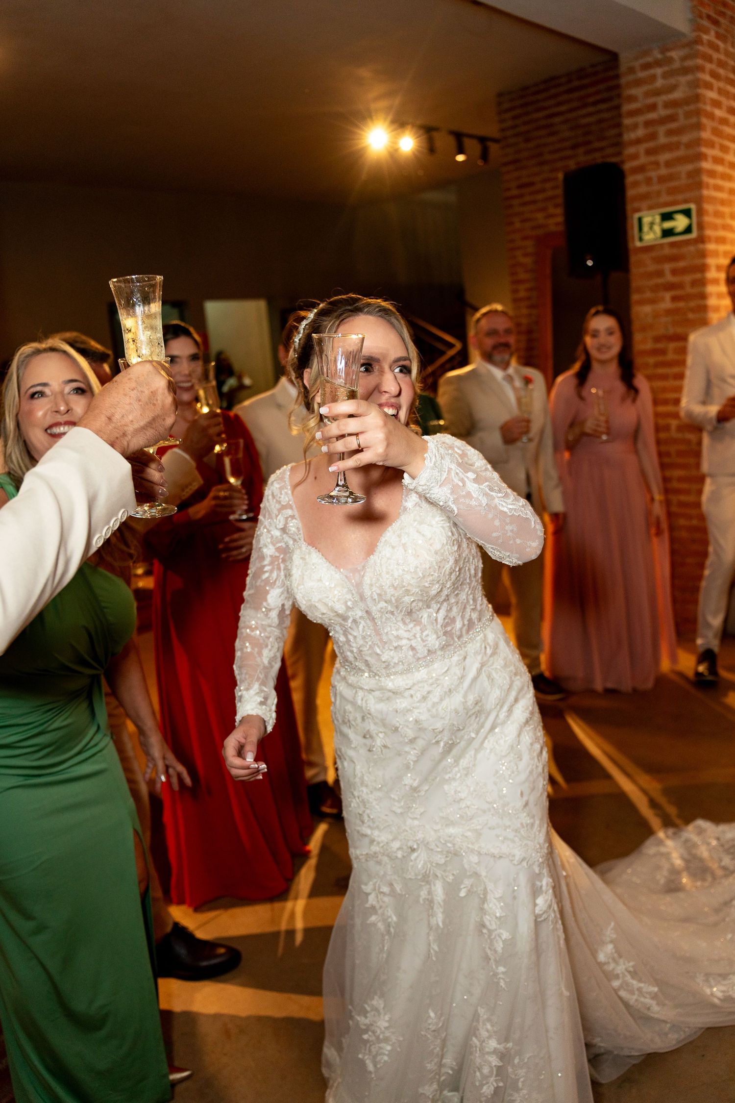 Bride in lace wedding gown raises champagne glass in toast surrounded by guests at wedding reception.