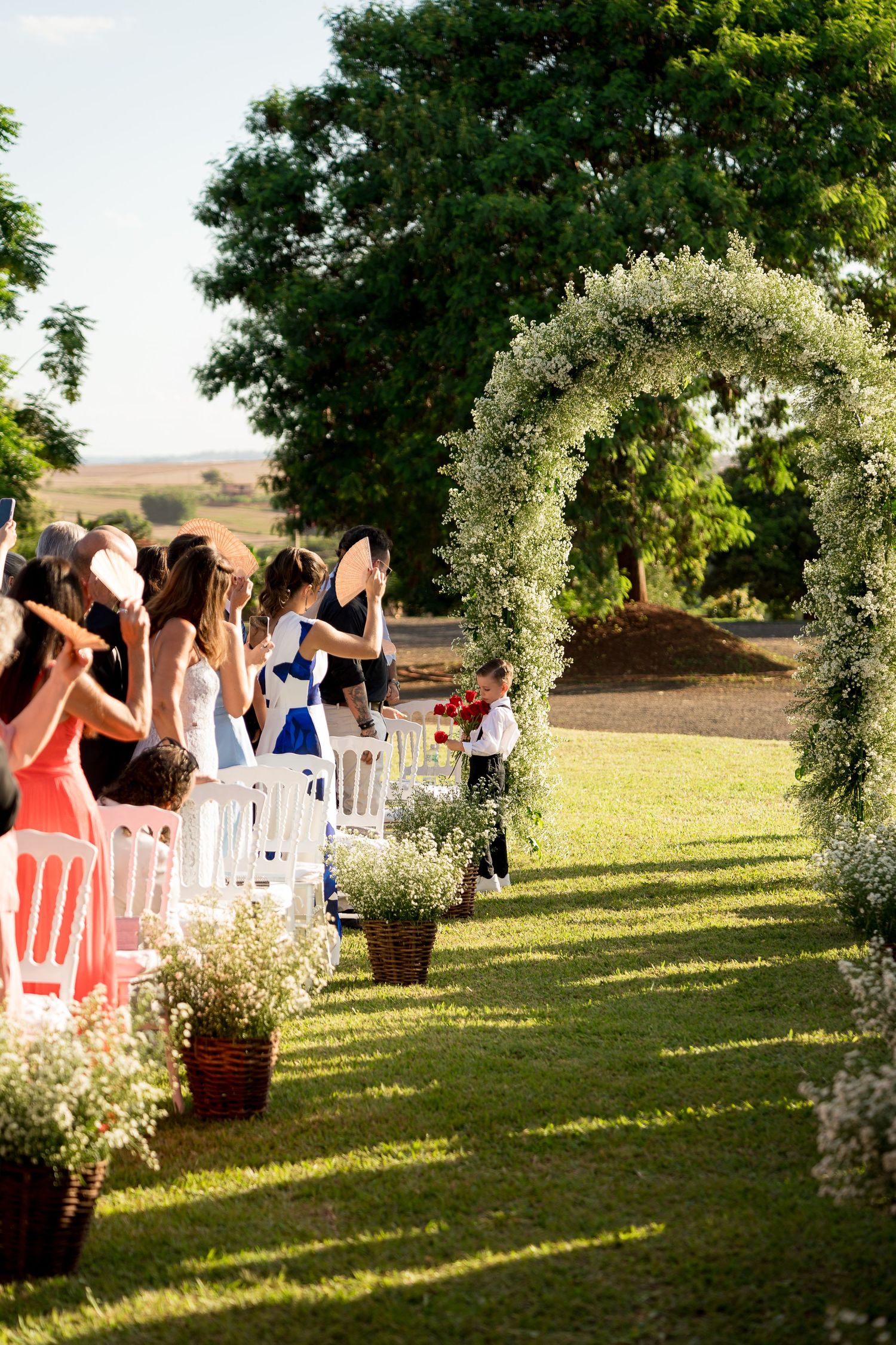Outdoor wedding ceremony with floral arch, white chairs, and baby's breath decorations on a sunny green lawn.