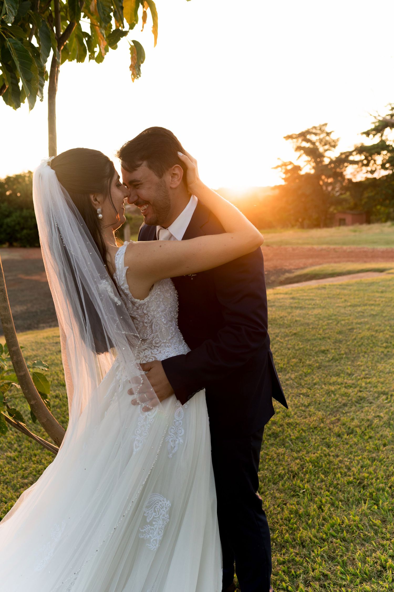 Bride and groom embrace at golden hour sunset in lush garden, wearing wedding dress with veil and black suit.