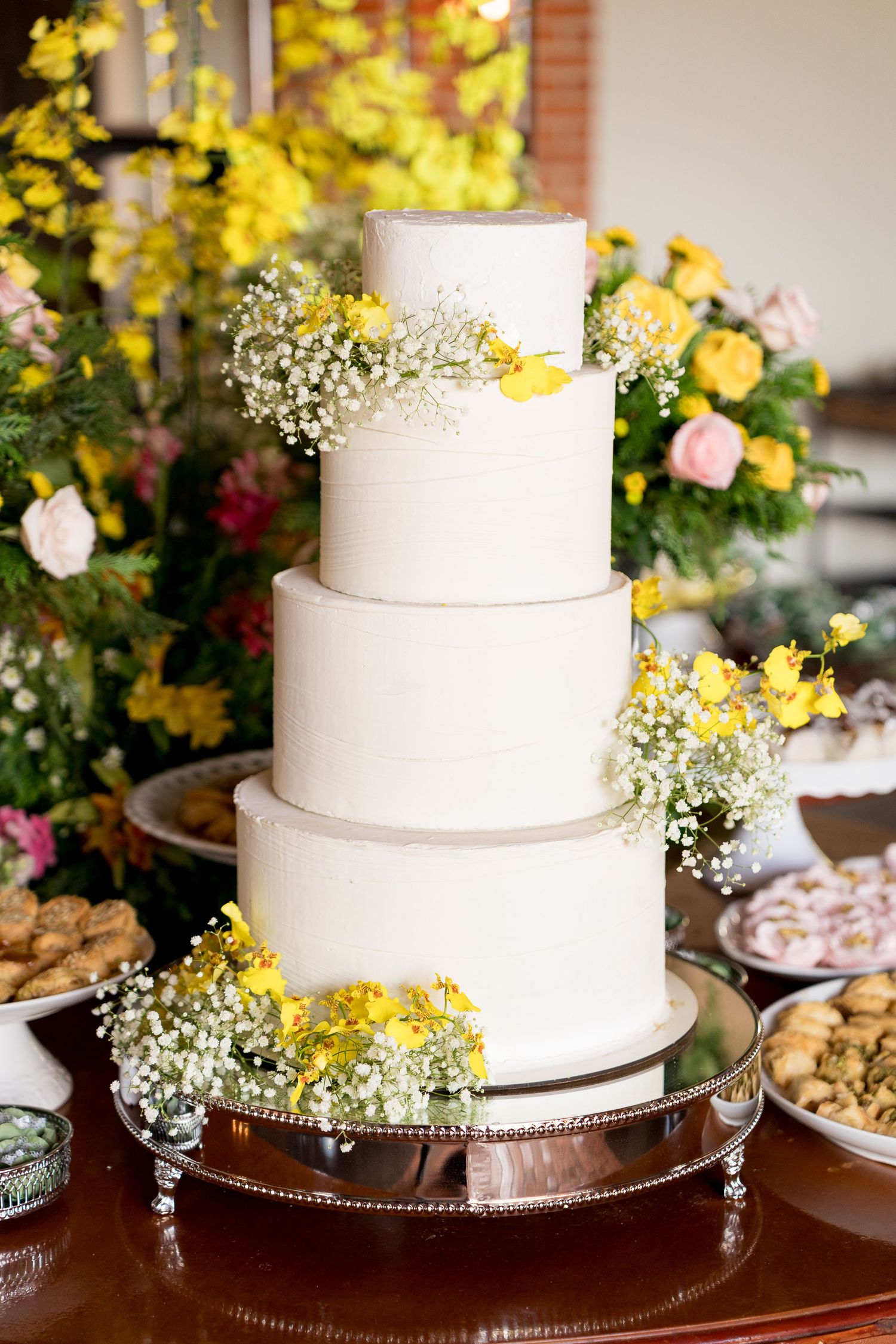Four-tier white wedding cake decorated with yellow flowers and baby's breath on a silver stand.