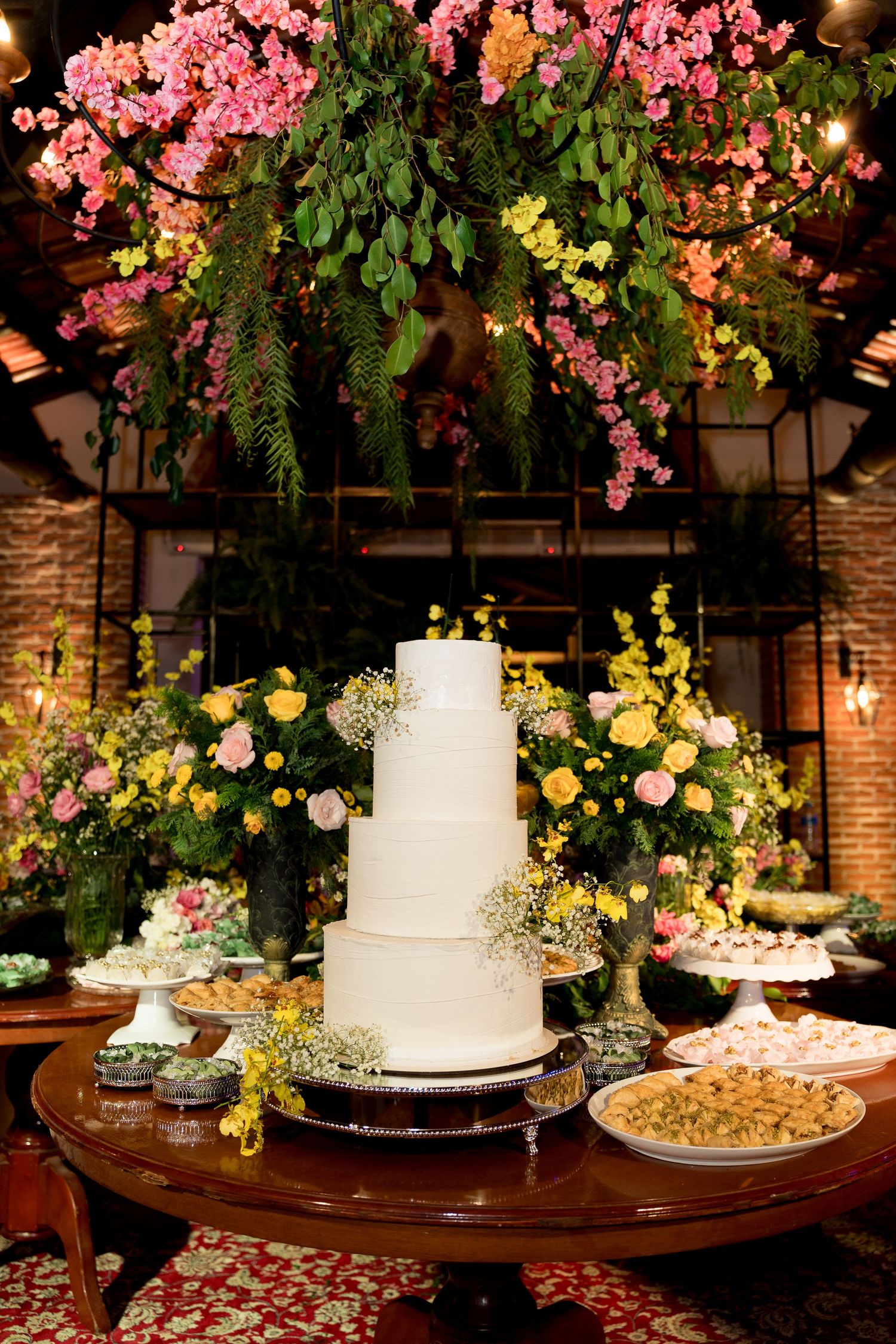 Elegant four-tier white wedding cake on wooden table surrounded by pink and yellow floral arrangements and desserts.