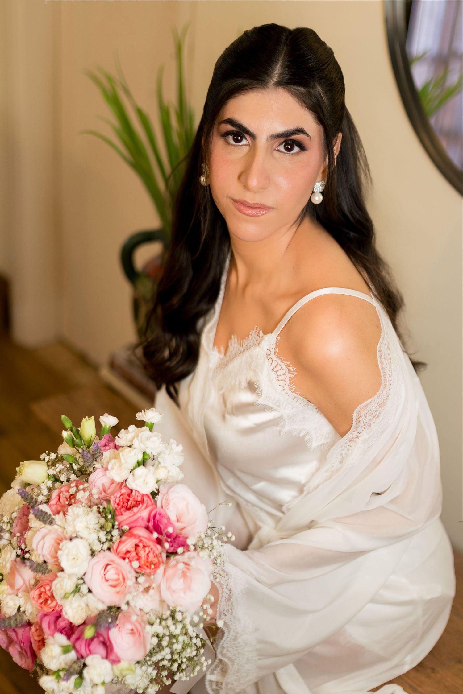 Bride in white satin robe holding pink and white floral bouquet, with pearl earrings and elegant makeup.