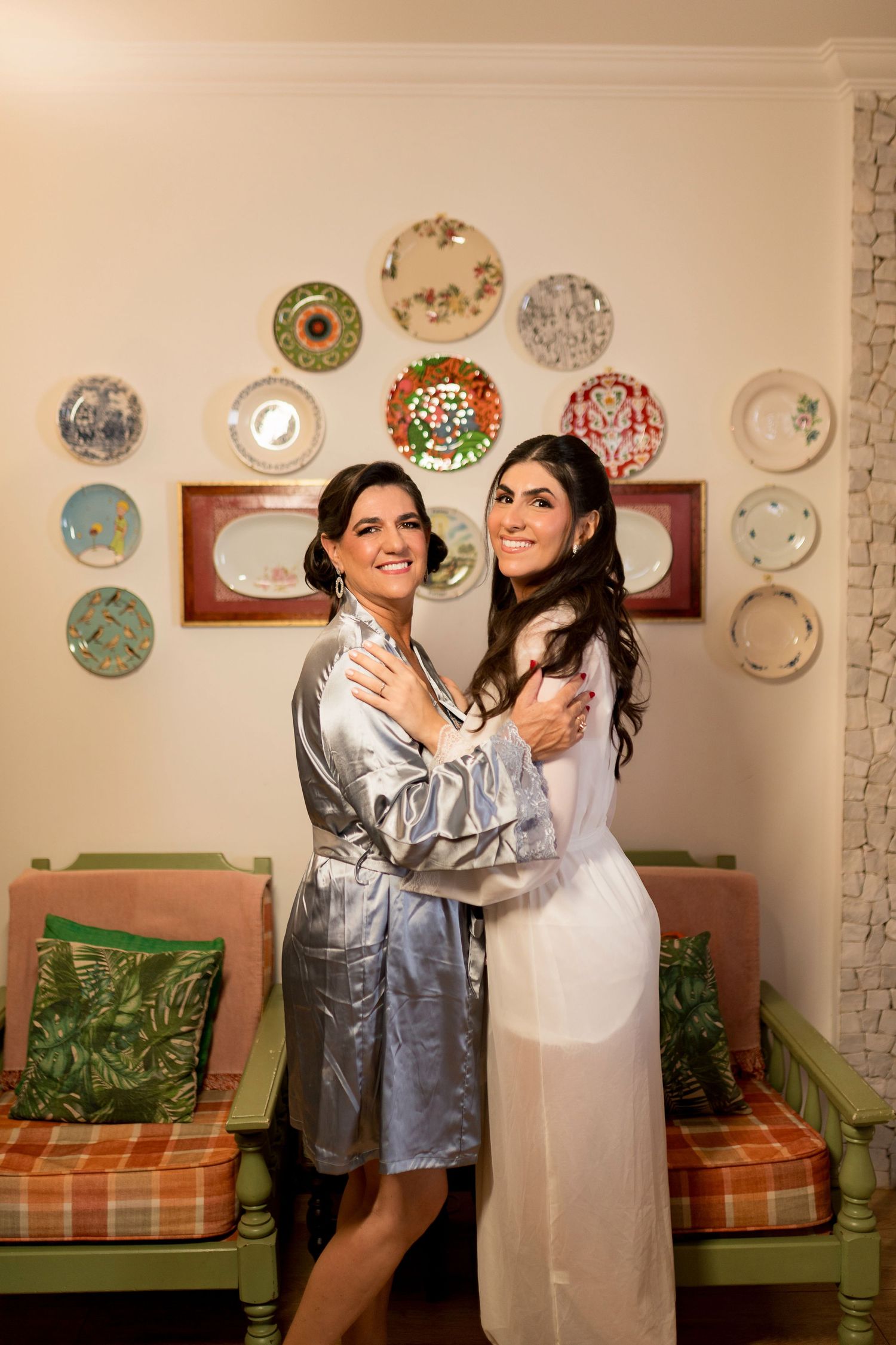 Two women in robes embrace smiling, standing before a decorative wall of plates in an elegant room.
