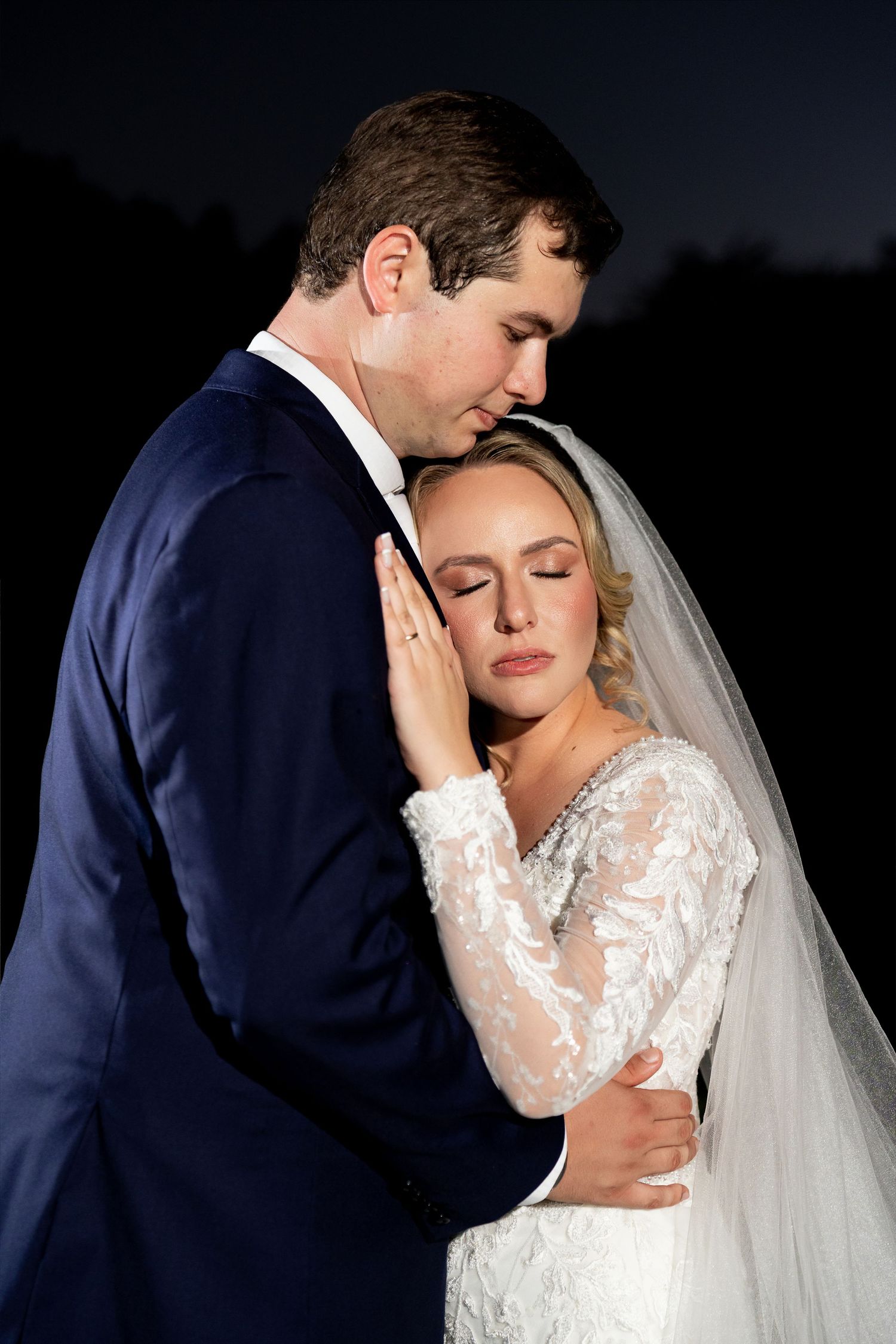 Bride and groom share tender embrace at night, groom kissing bride's head as she closes her eyes in lace wedding gown.