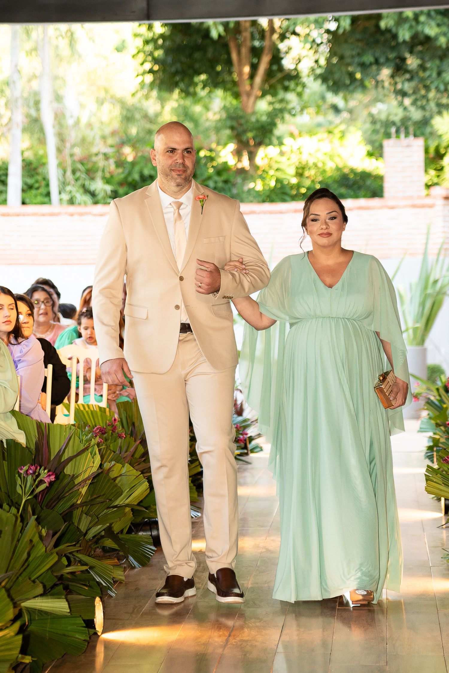 A man in a beige suit escorts a woman in a mint green gown down a wedding aisle outdoors.