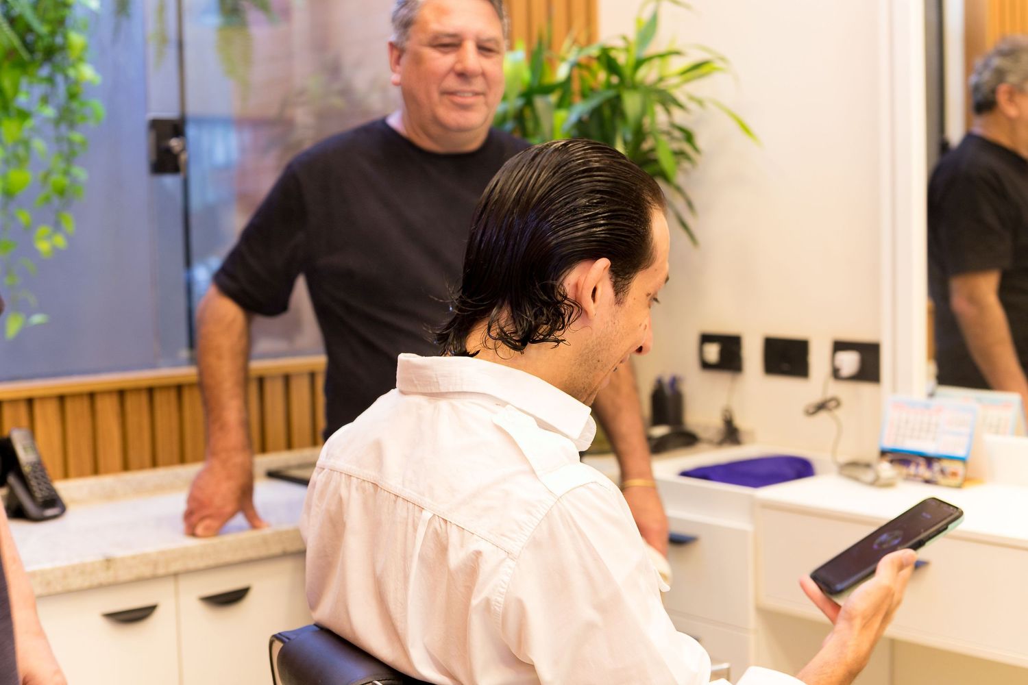 A man sits in a barber chair looking at his phone while a smiling barber stands behind him in a modern salon.