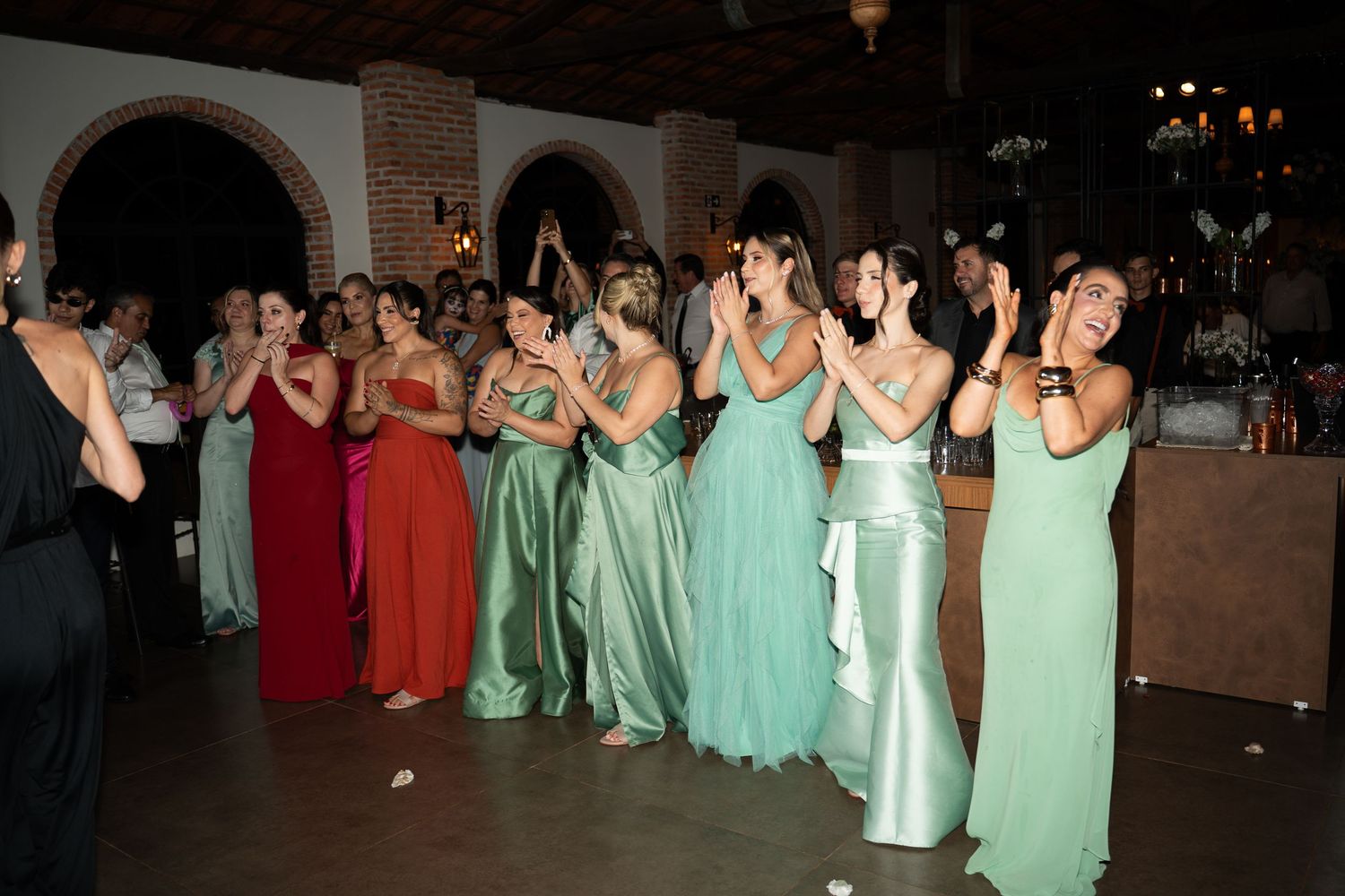 Women in elegant green and red gowns clapping at a formal event or wedding reception indoors.