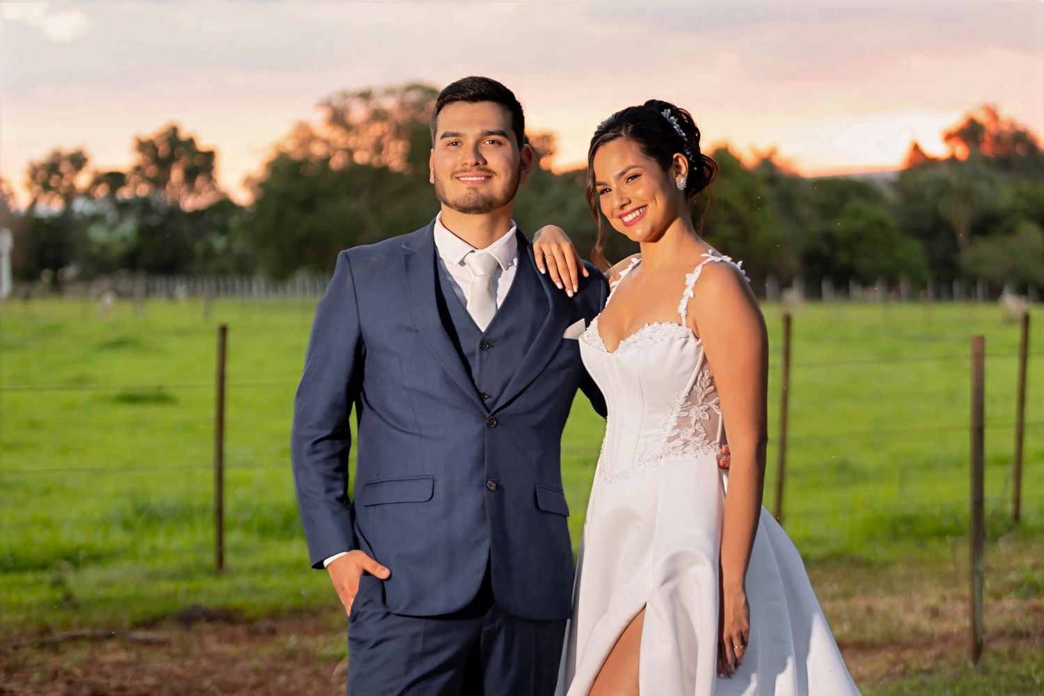 A couple in wedding attire poses outdoors at sunset, man in blue suit and woman in white bridal gown on green field.
