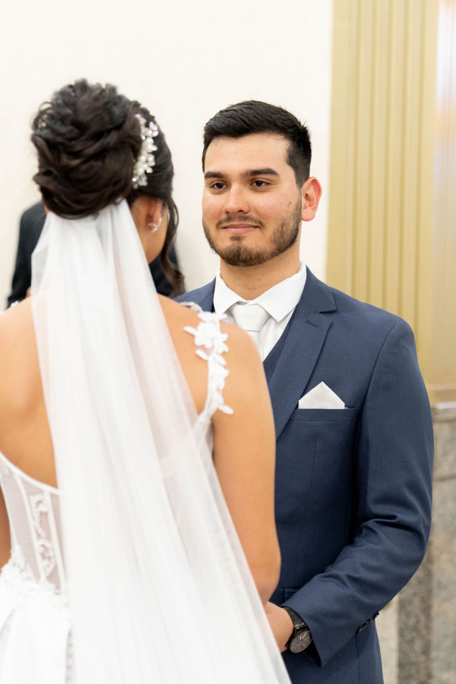 Groom in navy suit smiling at bride in white lace wedding dress and veil during wedding ceremony.