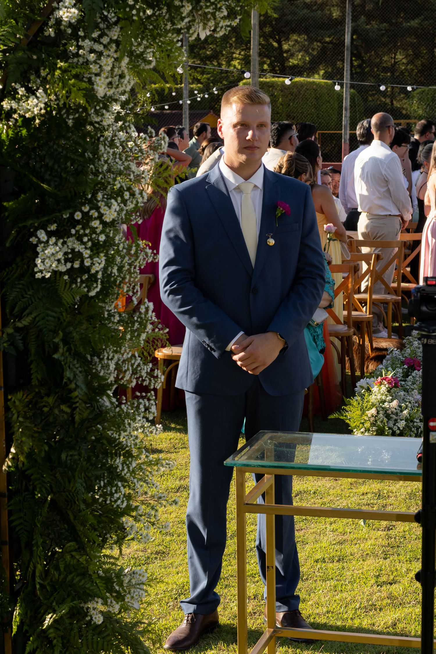Young man in blue suit with boutonniere stands at outdoor wedding ceremony beside floral arch on green lawn.