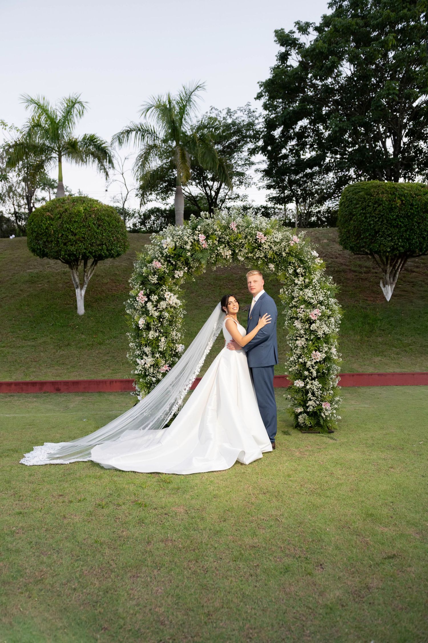 Bride and groom posing under a floral arch on a green lawn, surrounded by palm trees at an outdoor wedding.