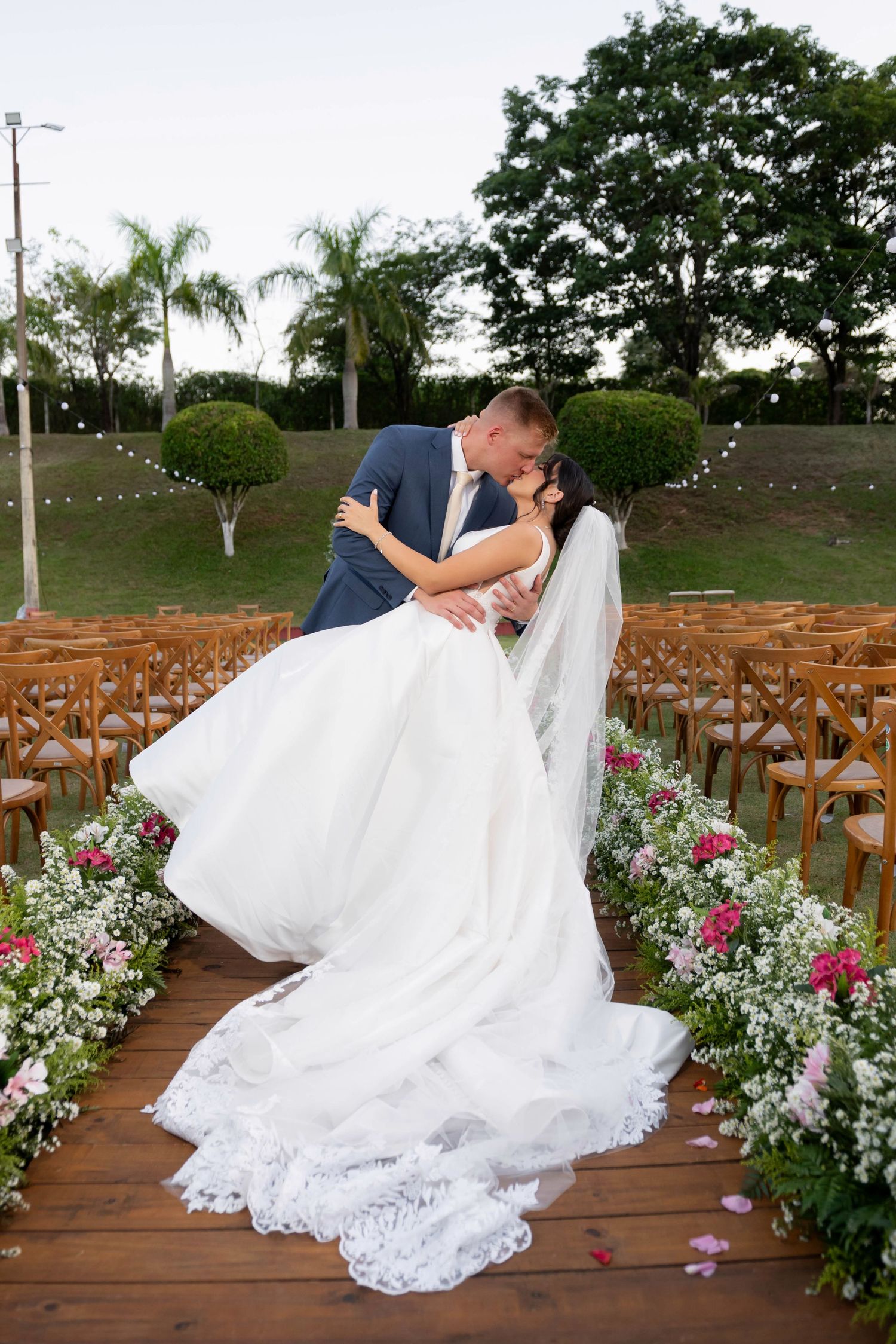 Bride and groom share a romantic dip kiss at outdoor wedding ceremony aisle lined with pink and white flowers.