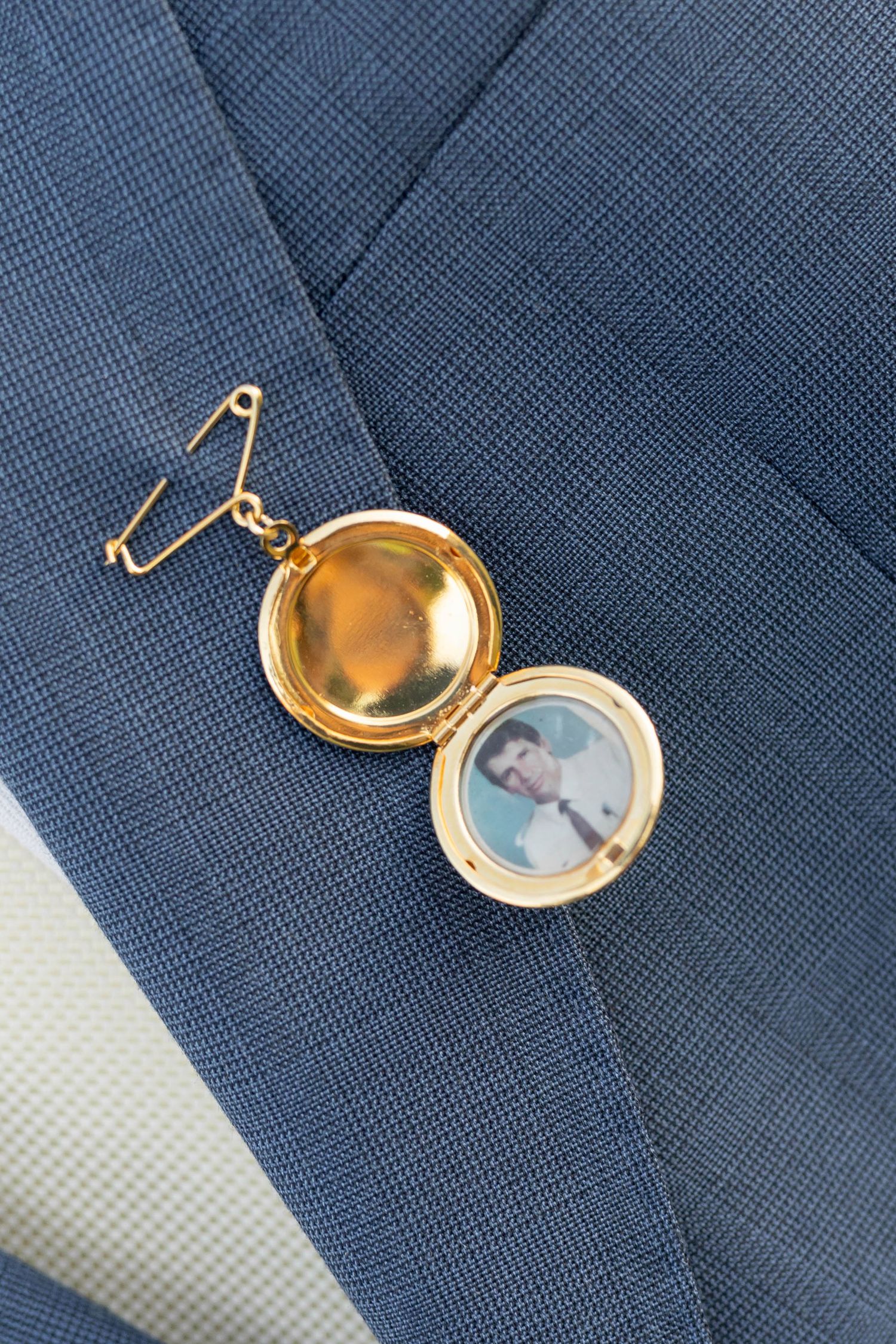 Gold locket pinned to blue suit lapel, open to reveal a small portrait photo inside, wedding detail shot.