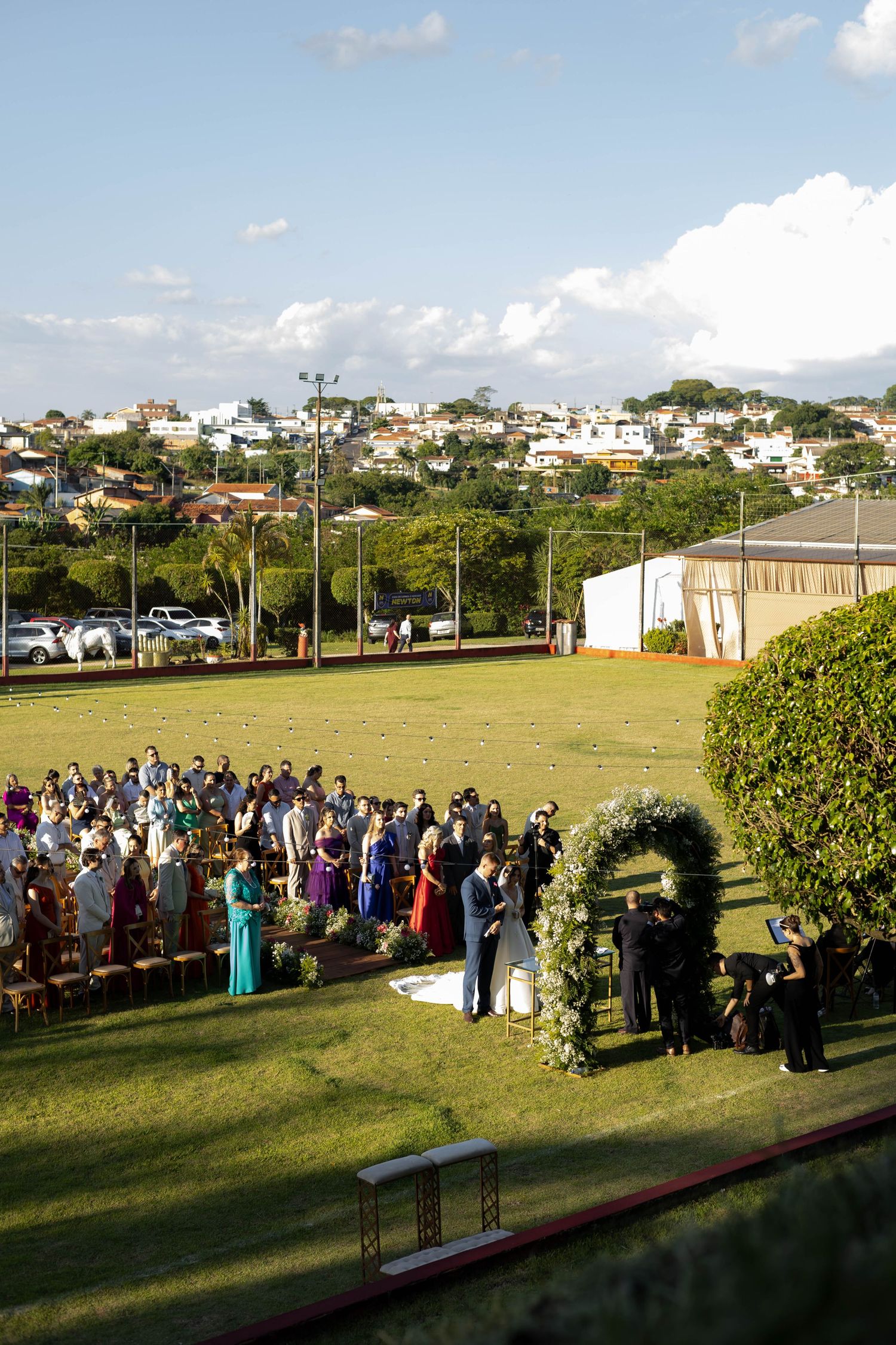 Outdoor wedding ceremony on green lawn with guests seated, floral arch, and hillside town in background.