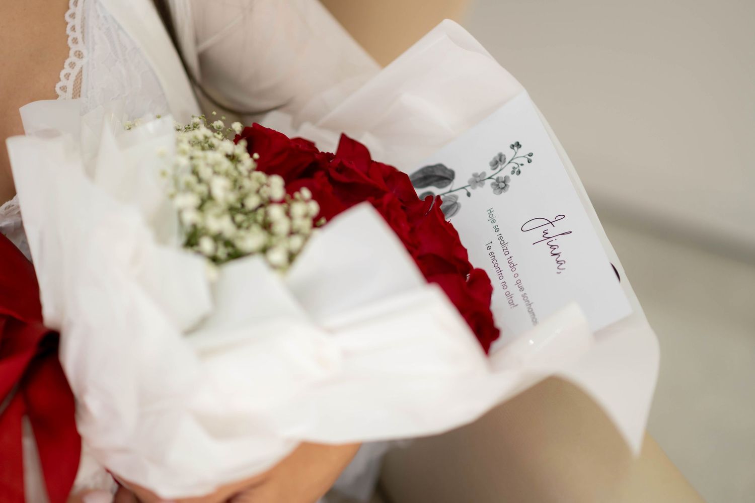 Close-up of a bridal bouquet with red roses, white baby's breath, and a decorative card wrapped in white tissue paper.