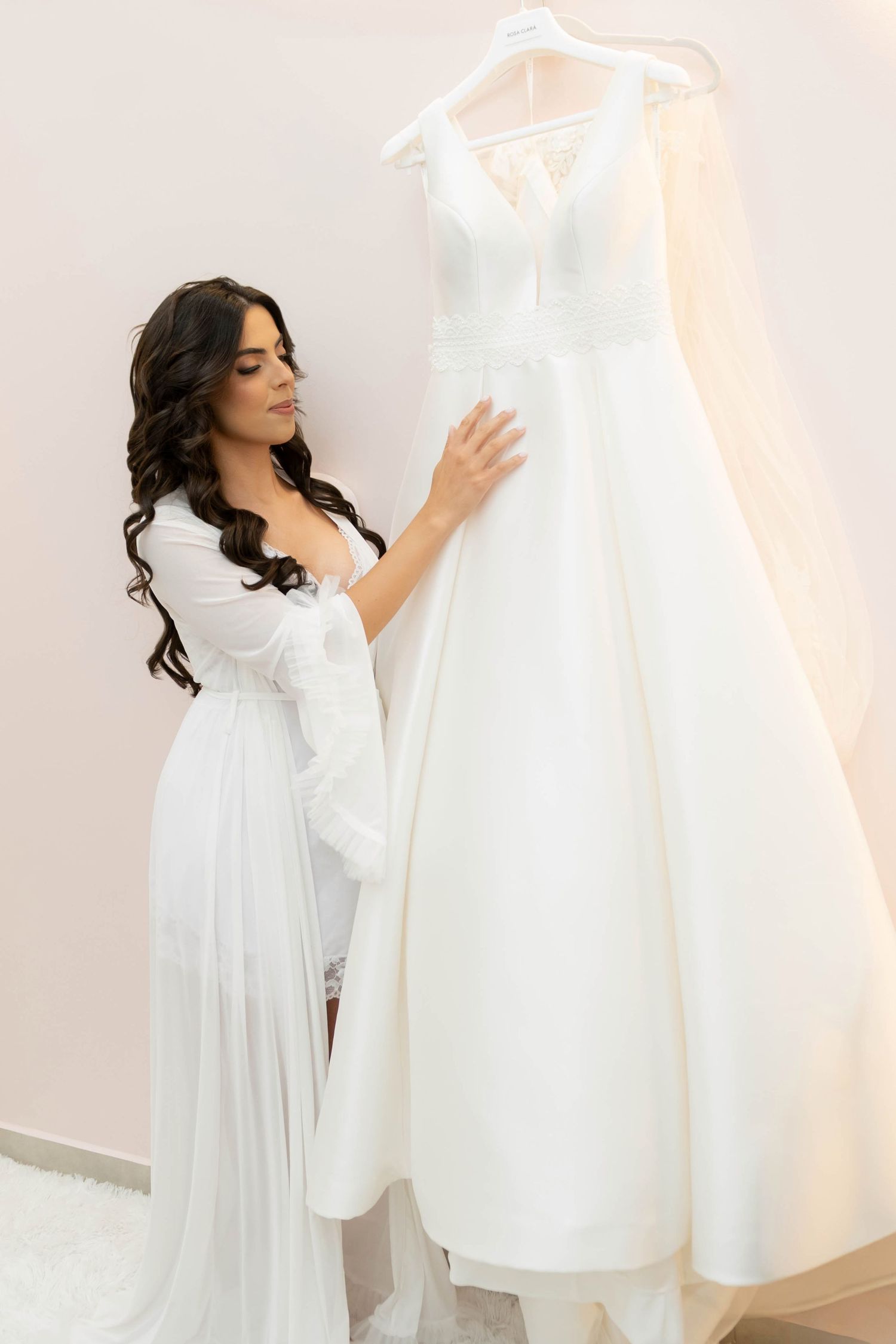 A beautiful dark-haired bride in a white robe admires her elegant wedding gown hanging on a rack in a bridal boutique.
