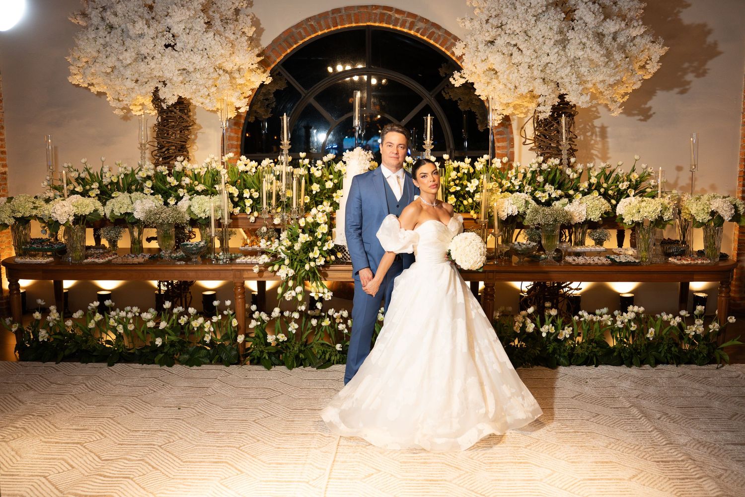 Bride and groom pose together on dance floor surrounded by lush white floral arrangements at elegant wedding reception.