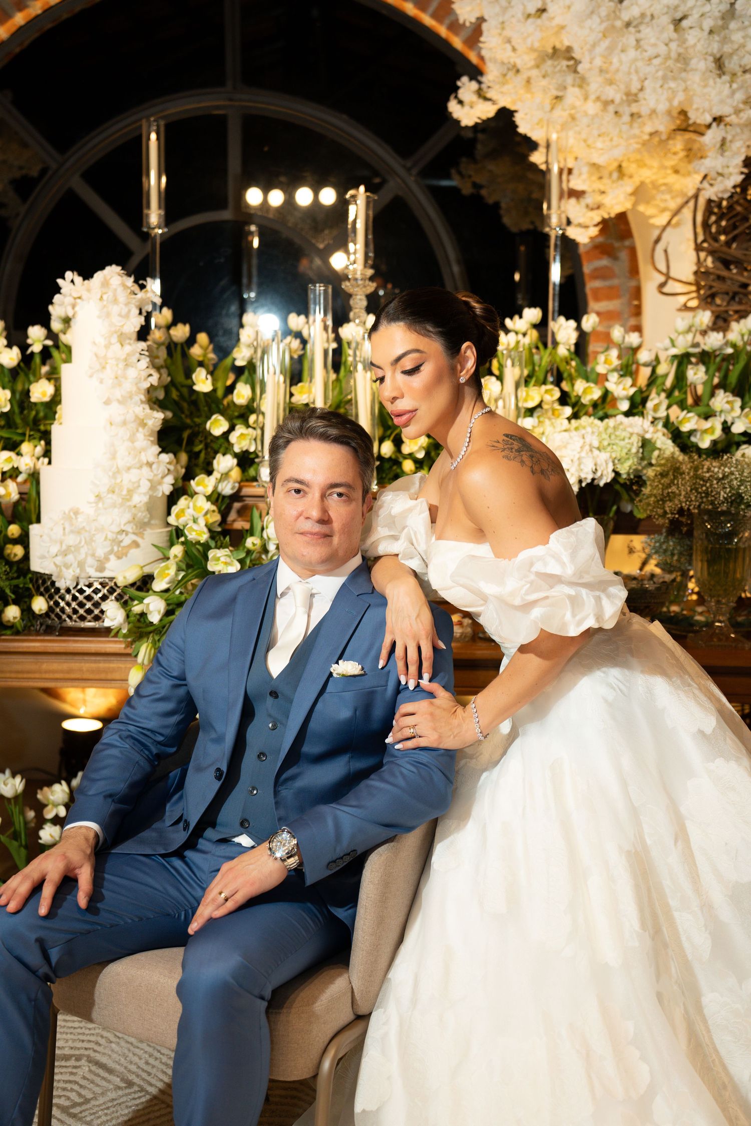 Elegant wedding couple posing before floral backdrop, groom in blue suit, bride in off-shoulder white gown with tattoo.