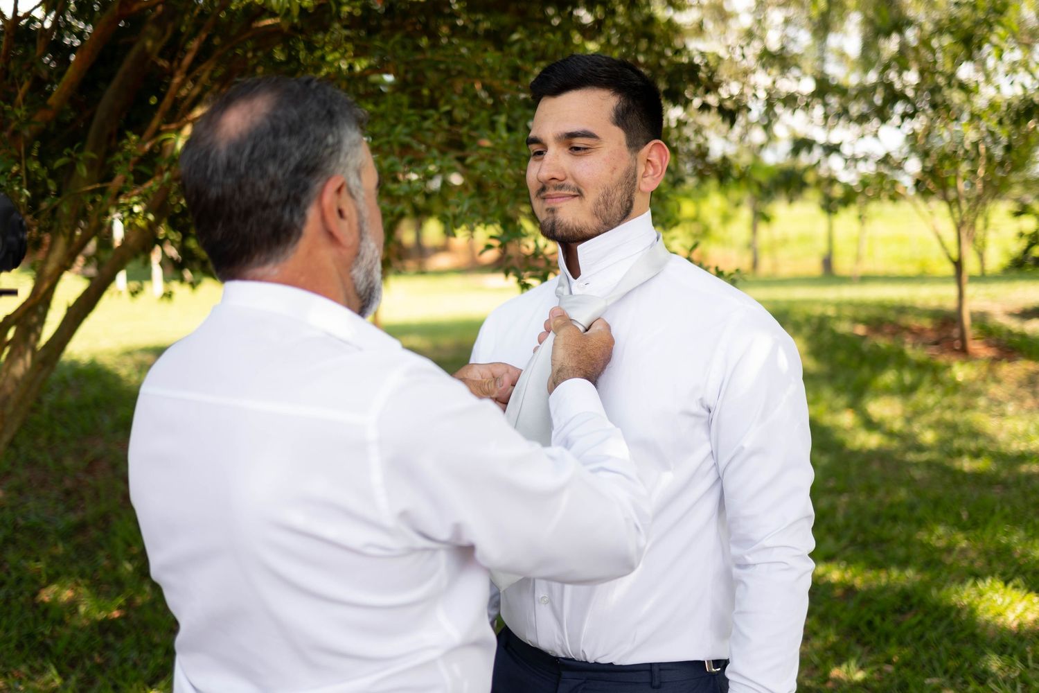 Older man adjusting white tie on young groom in white shirt outdoors before wedding ceremony.