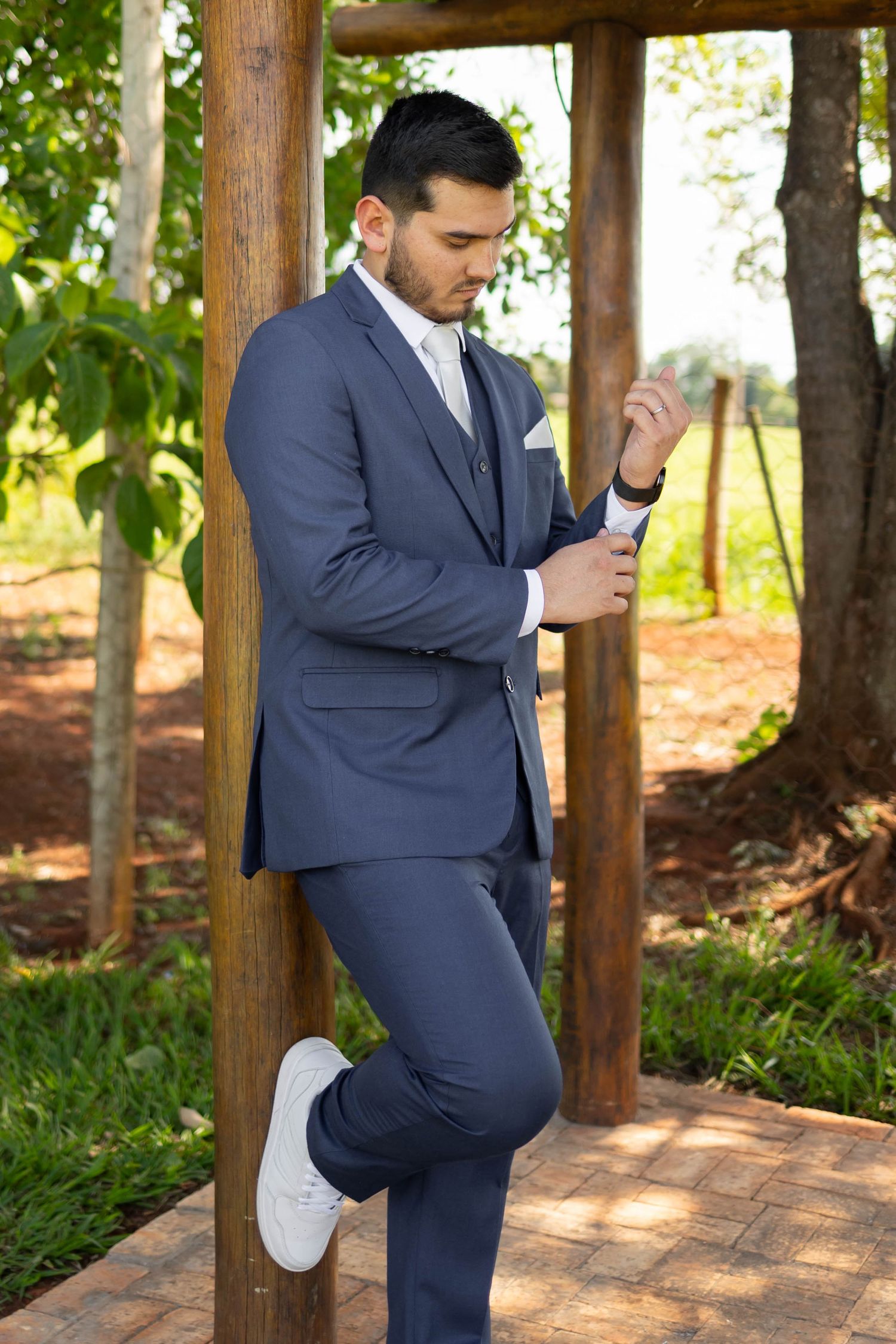 Young man in navy blue three-piece suit adjusting cufflinks outdoors, leaning against wooden post with white sneakers.