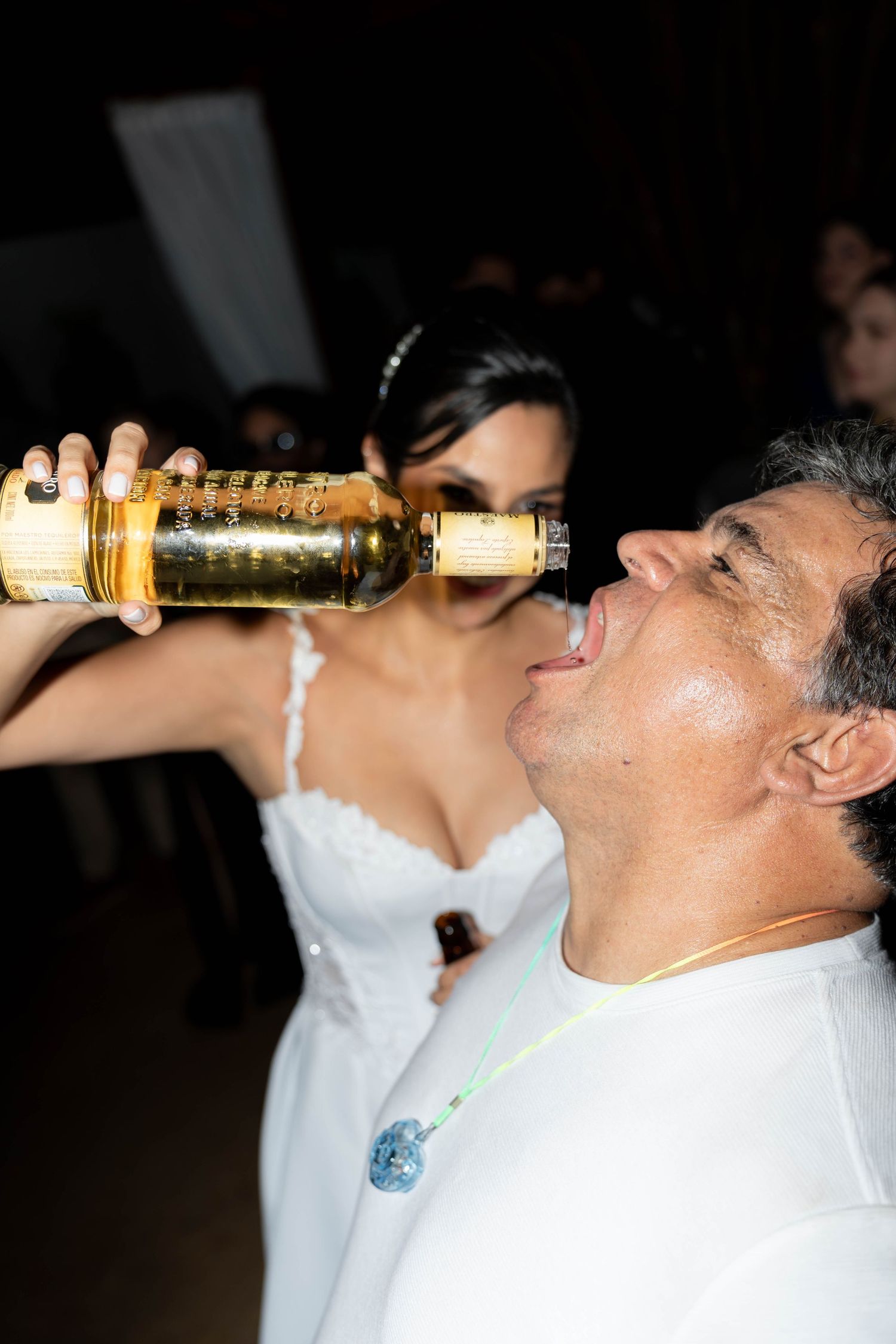 Bride in white dress pours whiskey bottle into groom's mouth at wedding reception celebration.