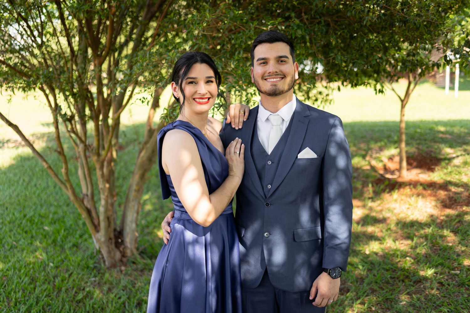 Couple in matching navy blue formal wear posing outdoors in front of green trees for a special occasion.