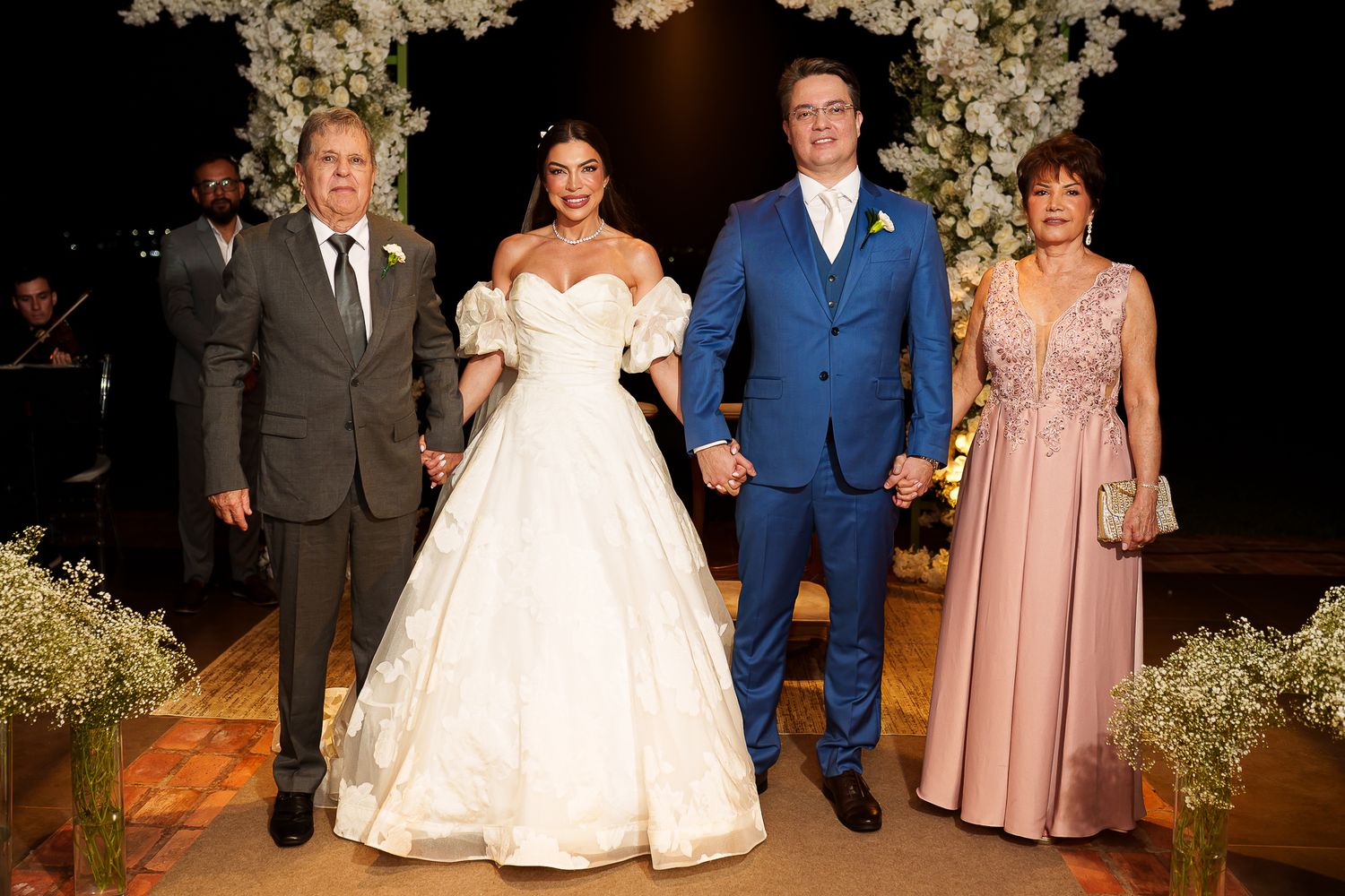 Bride in white gown and groom in blue suit pose with family members at elegant floral wedding ceremony.