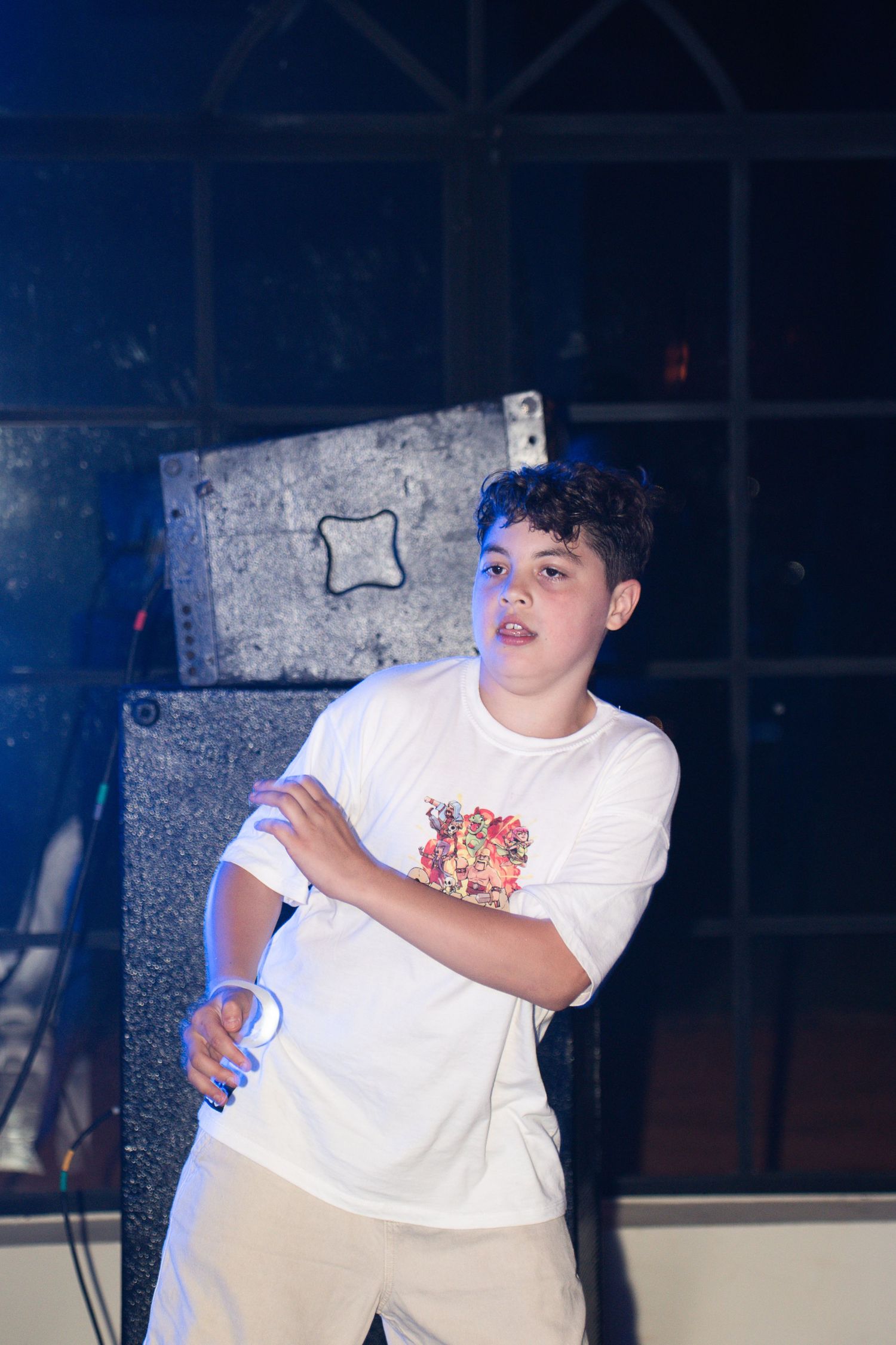 Young teen boy in white graphic t-shirt dancing on stage with speaker behind him at an indoor event.