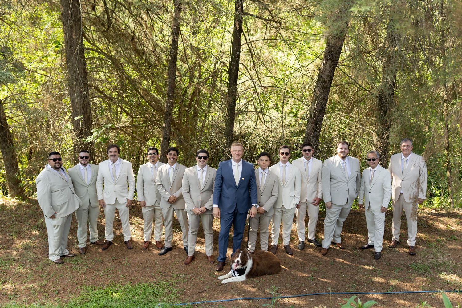 Groom in navy suit stands with groomsmen in tan suits and sunglasses in a forest wedding party photo.