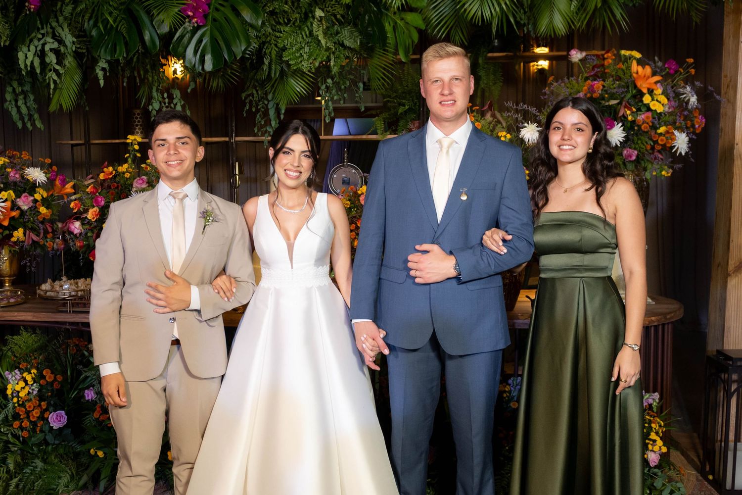 Wedding party of four posing elegantly, bride in white gown, groom in blue suit, guests in tan and green formal attire.