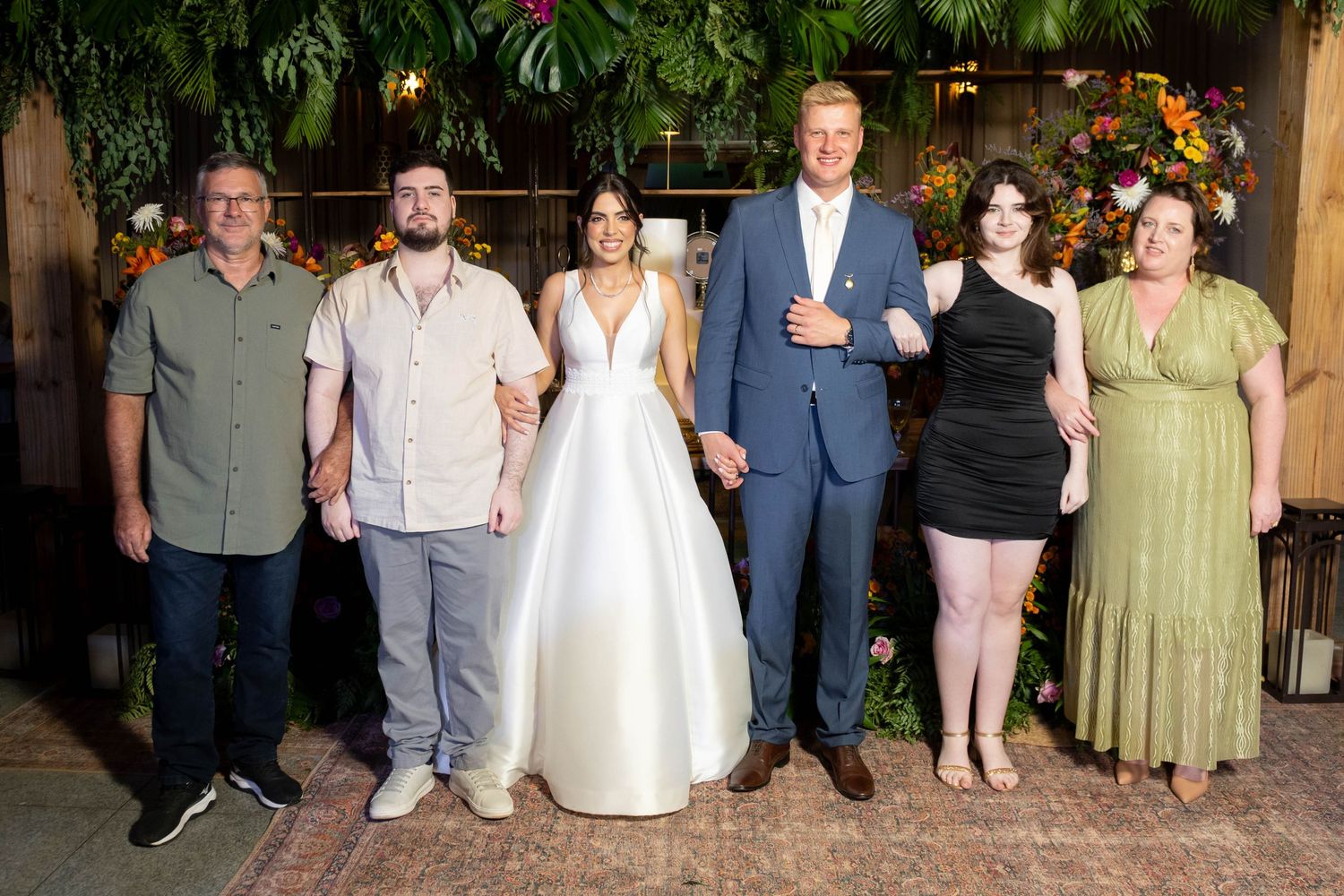 Bride and groom smile with four family members before colorful floral wall at wedding celebration.