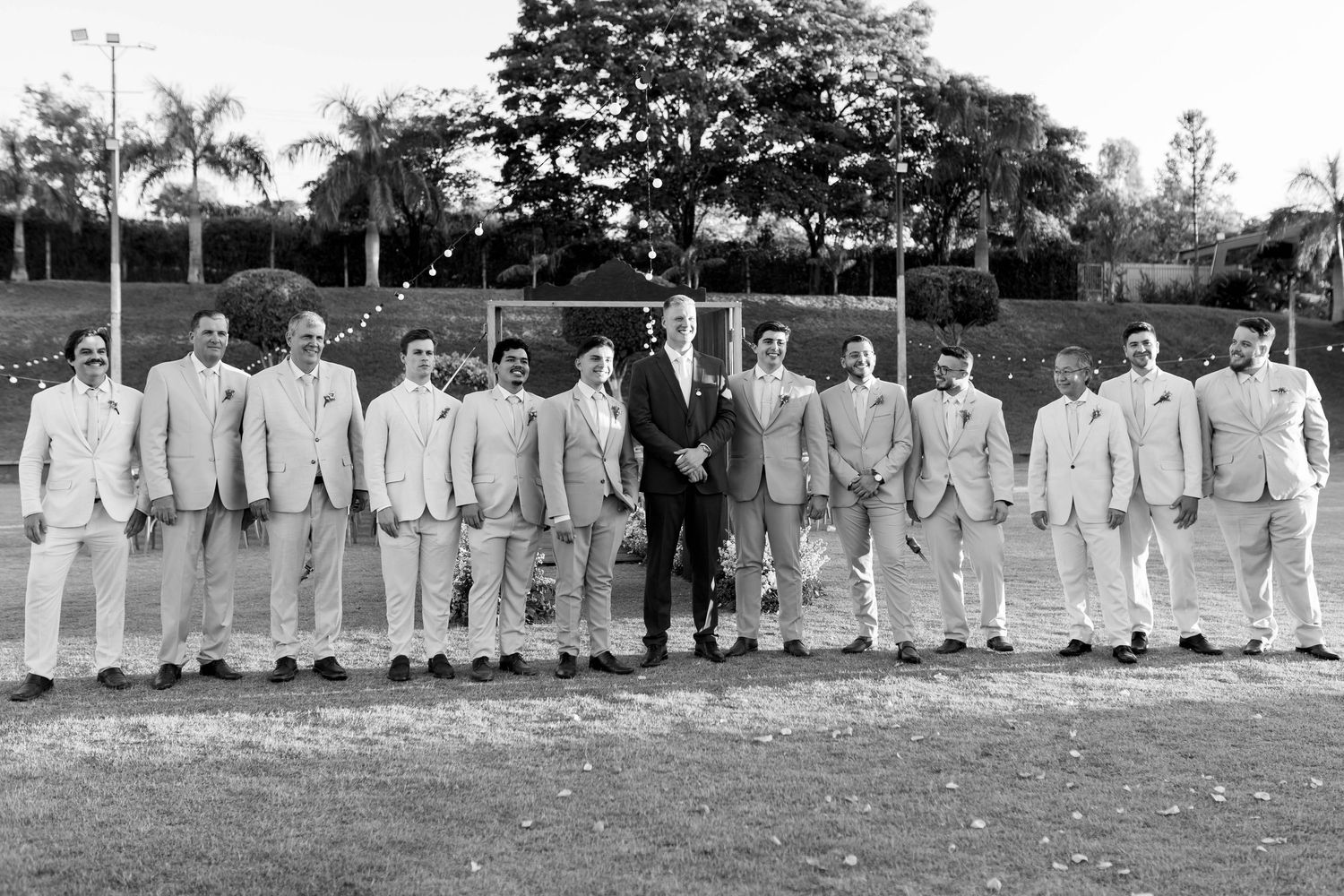 Groom and groomsmen in matching light suits posing together outdoors on a lawn at a wedding celebration.