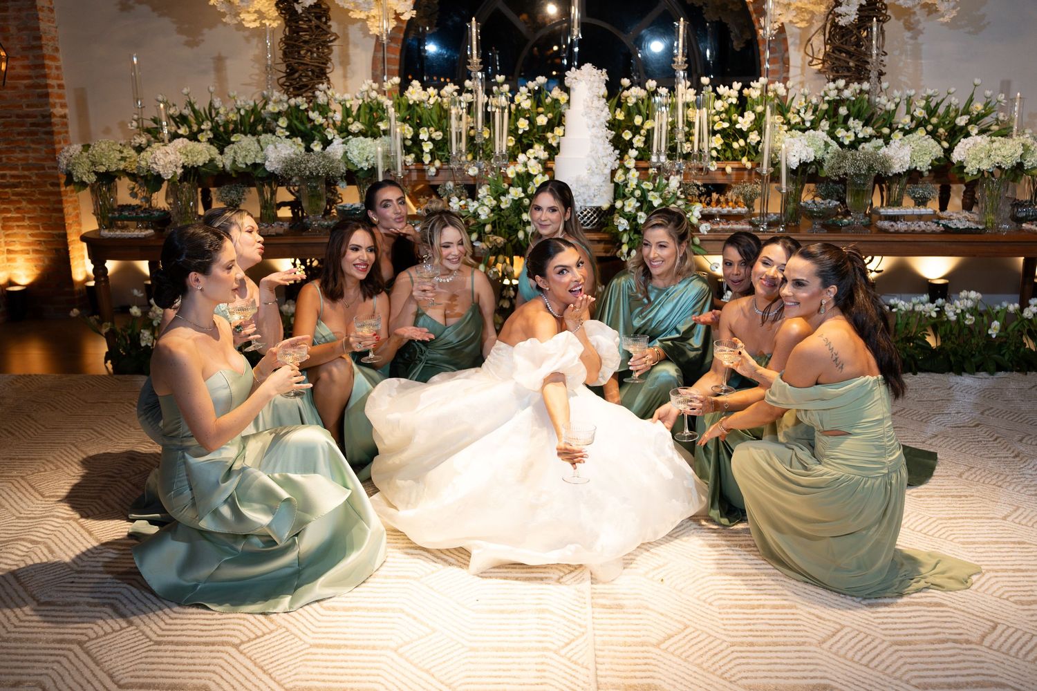 Bride in white gown sits on dance floor laughing with bridesmaids in sage green dresses at elegant floral wedding reception.