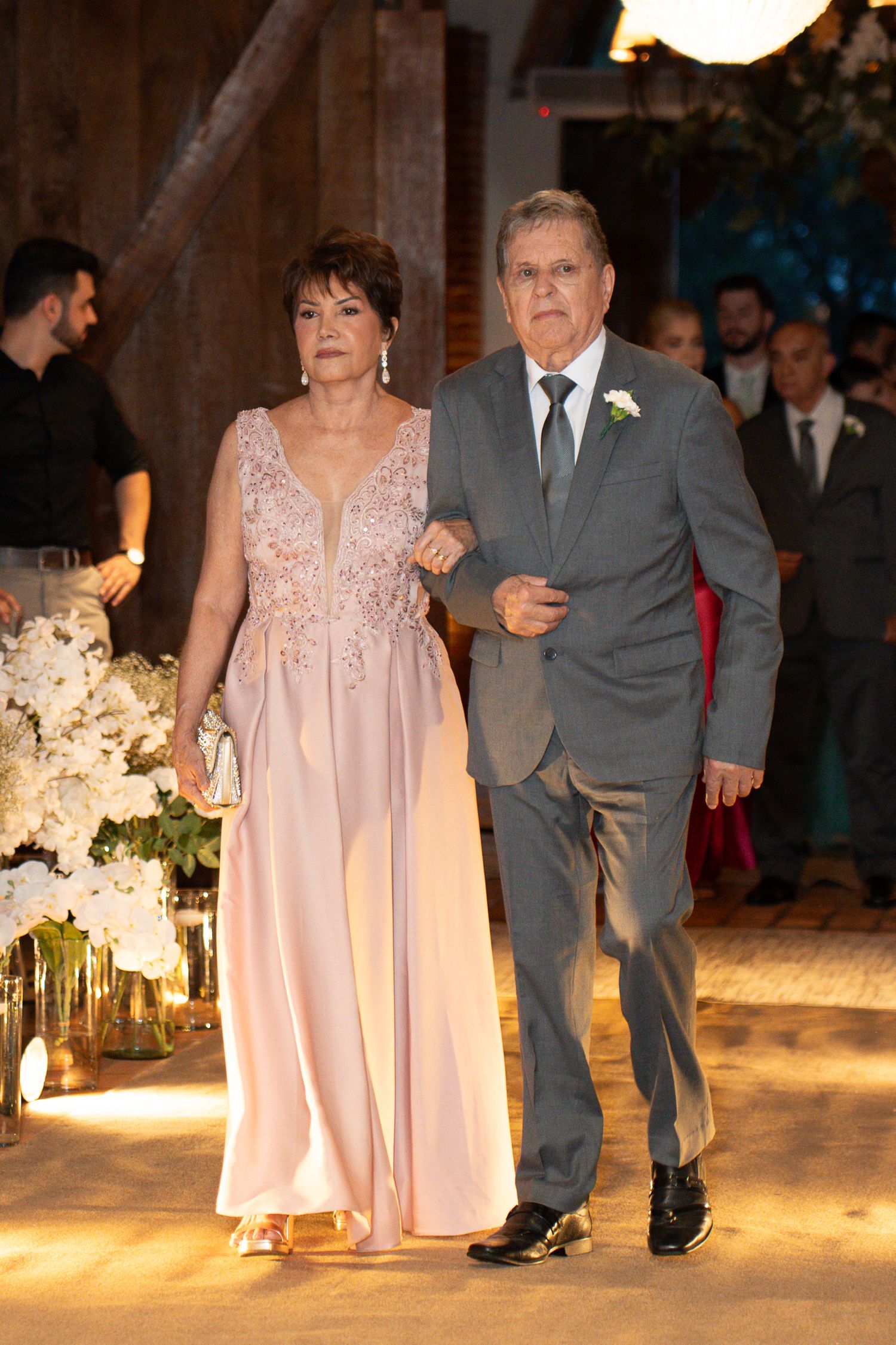 Elegant couple walking together at a formal wedding event, woman in pink gown, man in gray suit with boutonniere.