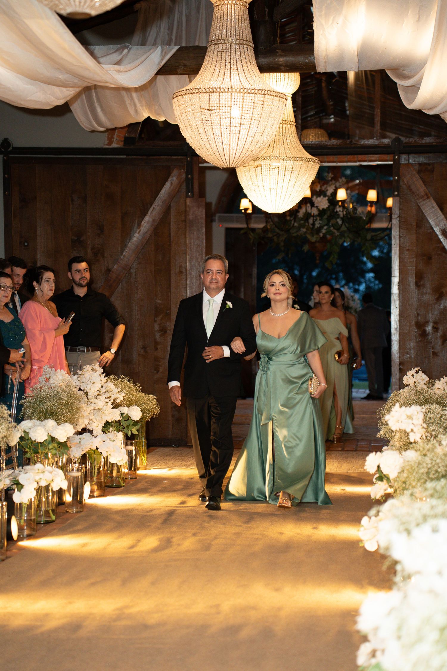 Couple walks down floral-lined aisle under crystal chandeliers in elegant barn wedding ceremony.