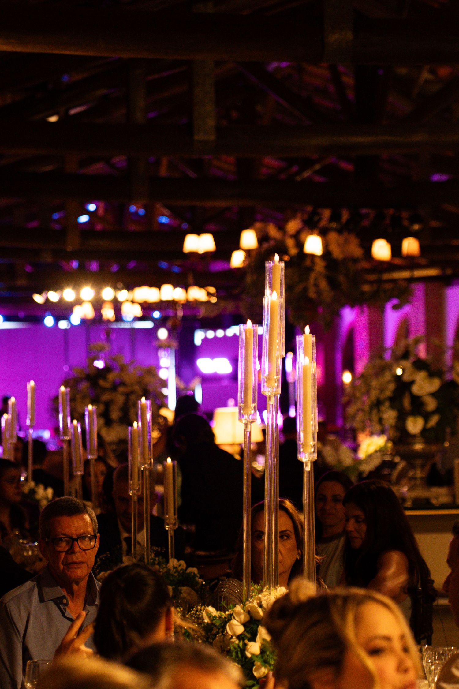 Elegant wedding reception with tall candelabras, glowing candles, floral arrangements, and purple ambient lighting in a rustic venue.