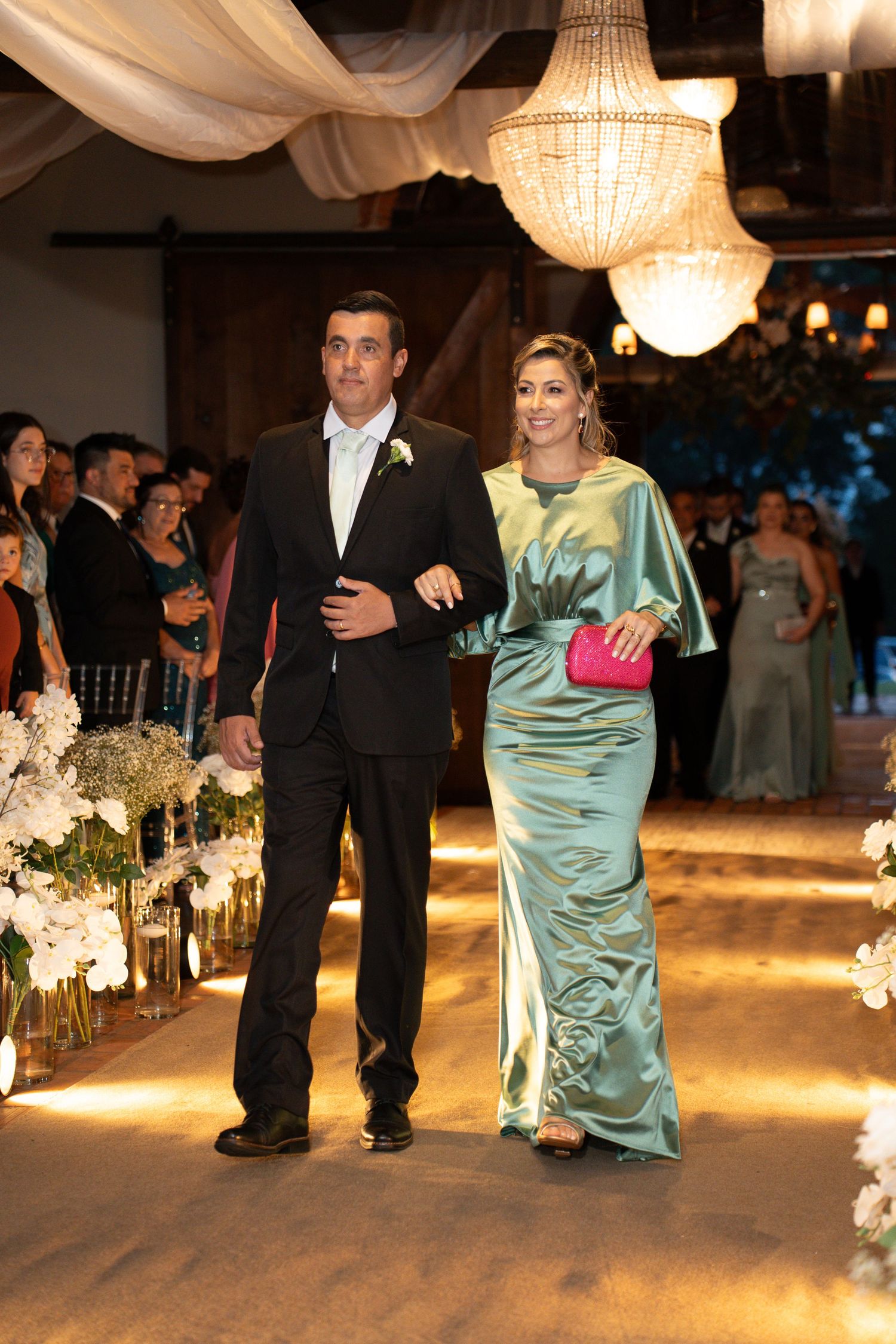 Elegant couple walking down wedding aisle, man in black suit, woman in teal satin gown with pink clutch, chandeliers above.