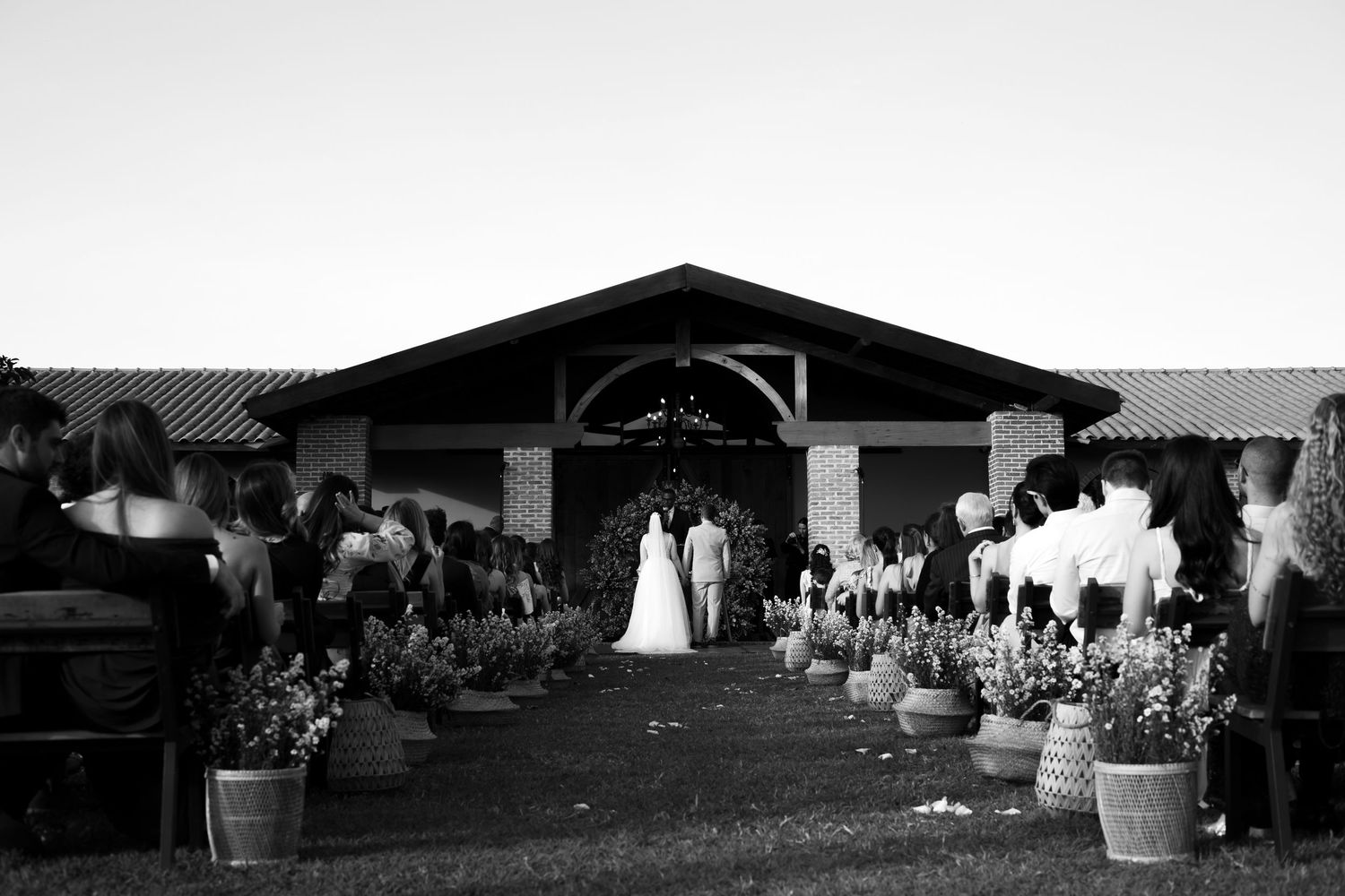 Black and white photo of a wedding ceremony outside a rustic brick venue with guests seated along a flower-lined aisle.