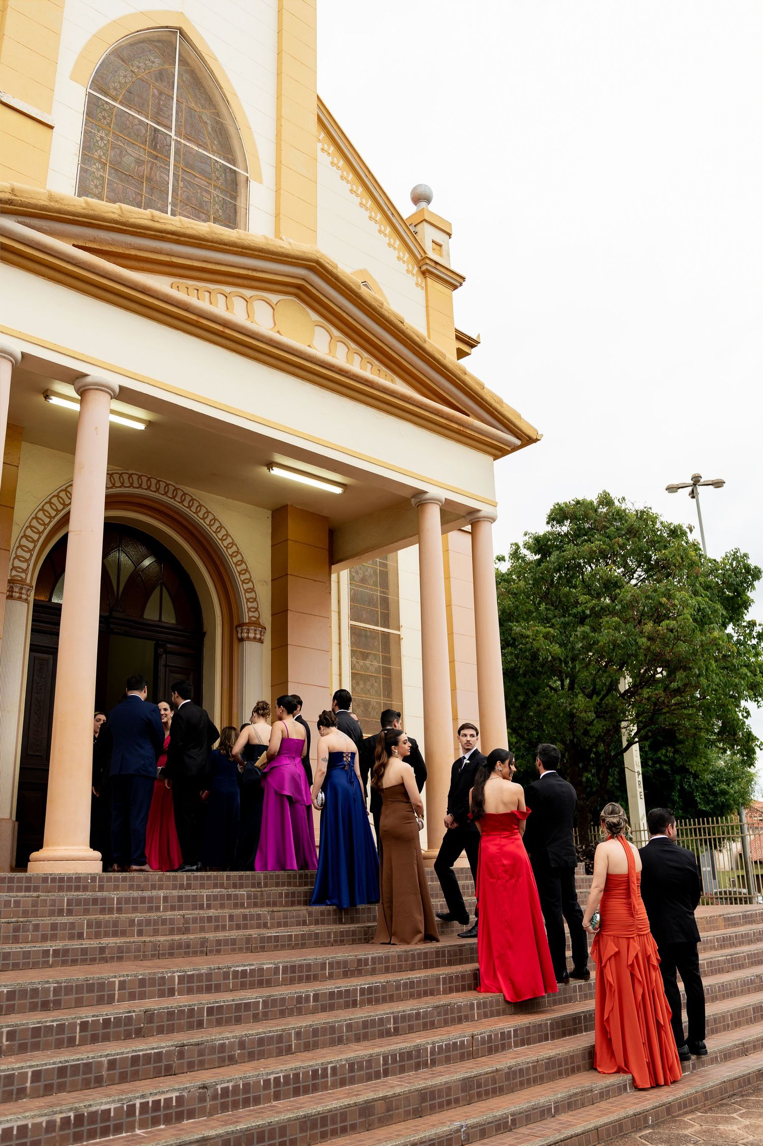 Elegantly dressed wedding guests in colorful gowns ascending brick steps of a yellow colonial church.