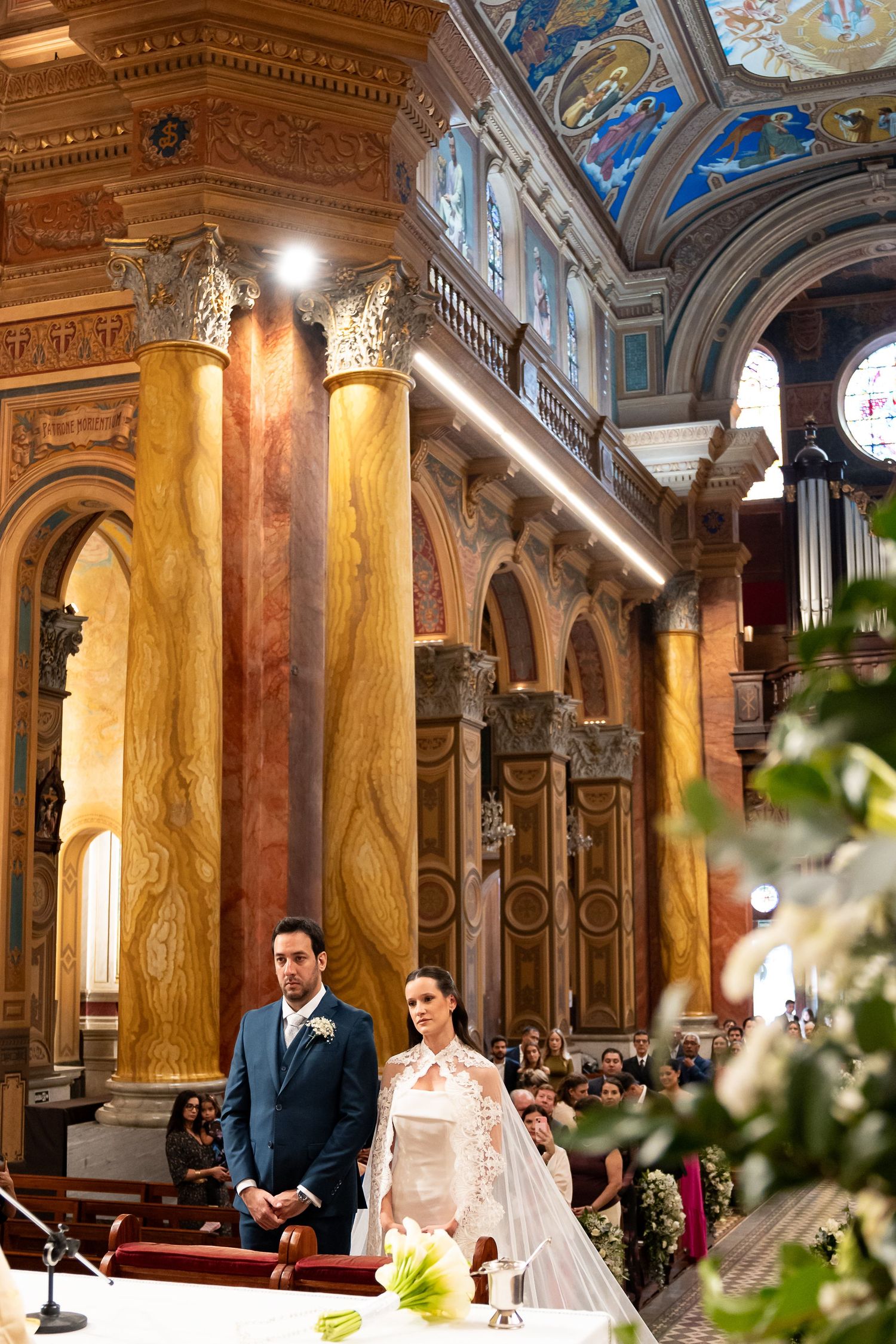 Bride and groom stand at the altar of an ornate church with golden columns and painted ceilings during wedding ceremony.