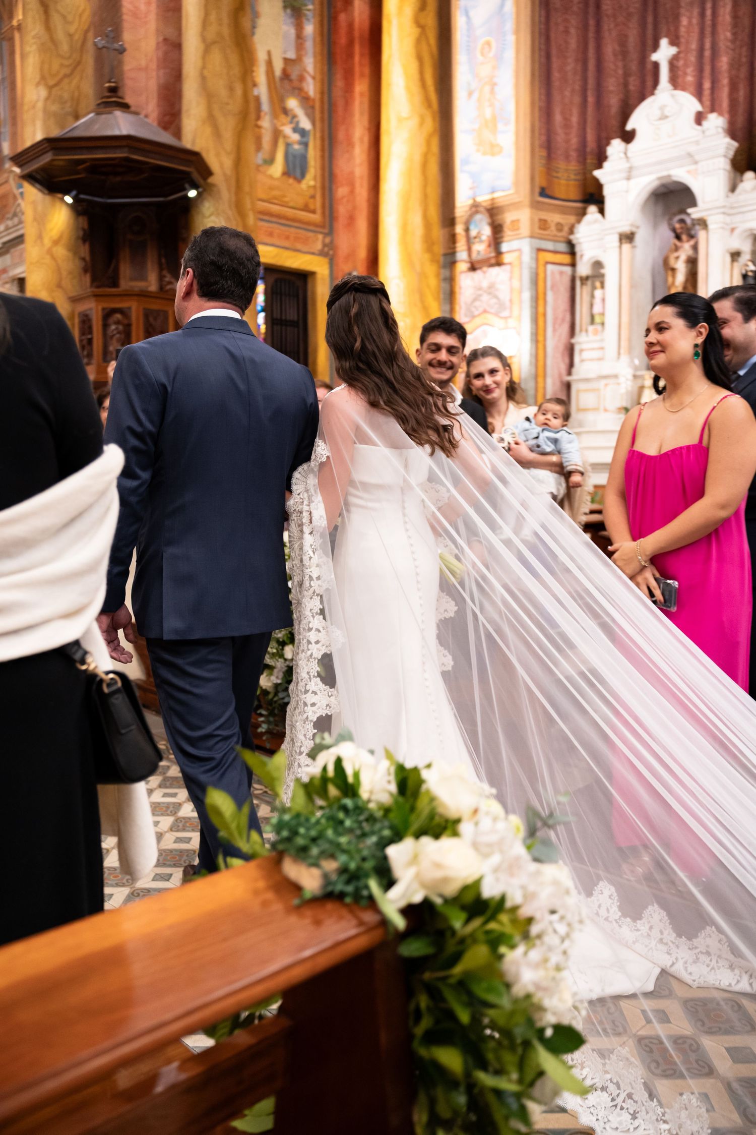 Bride in white gown with lace veil walks down church aisle with groom in navy suit during wedding ceremony.