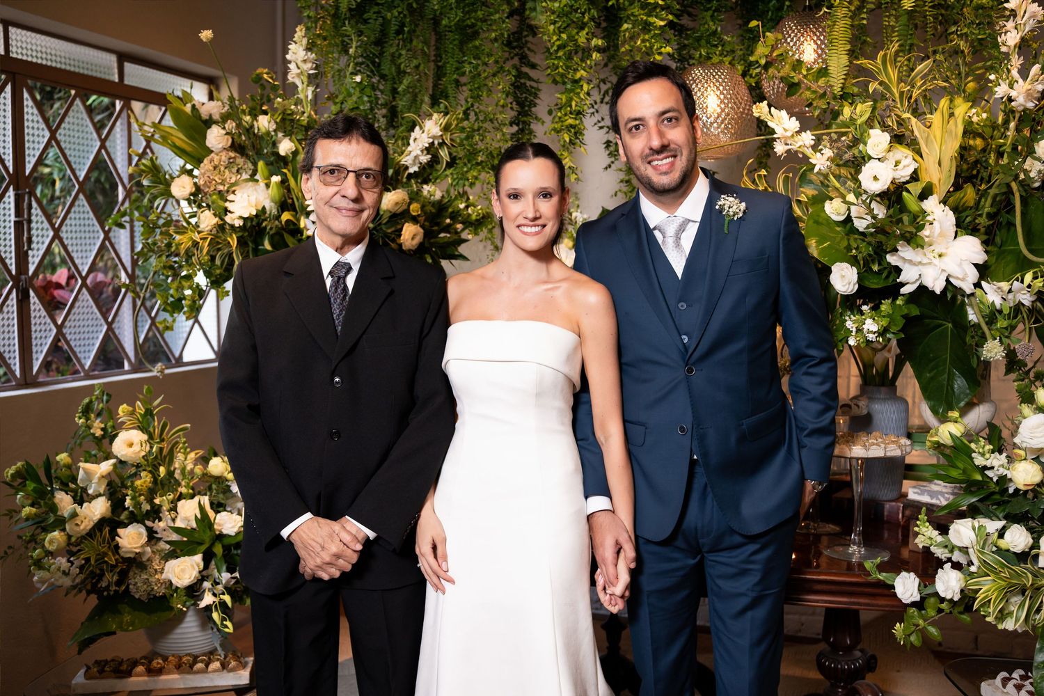 Bride and groom pose with officiant before lush floral arch of white flowers and greenery at elegant wedding ceremony.
